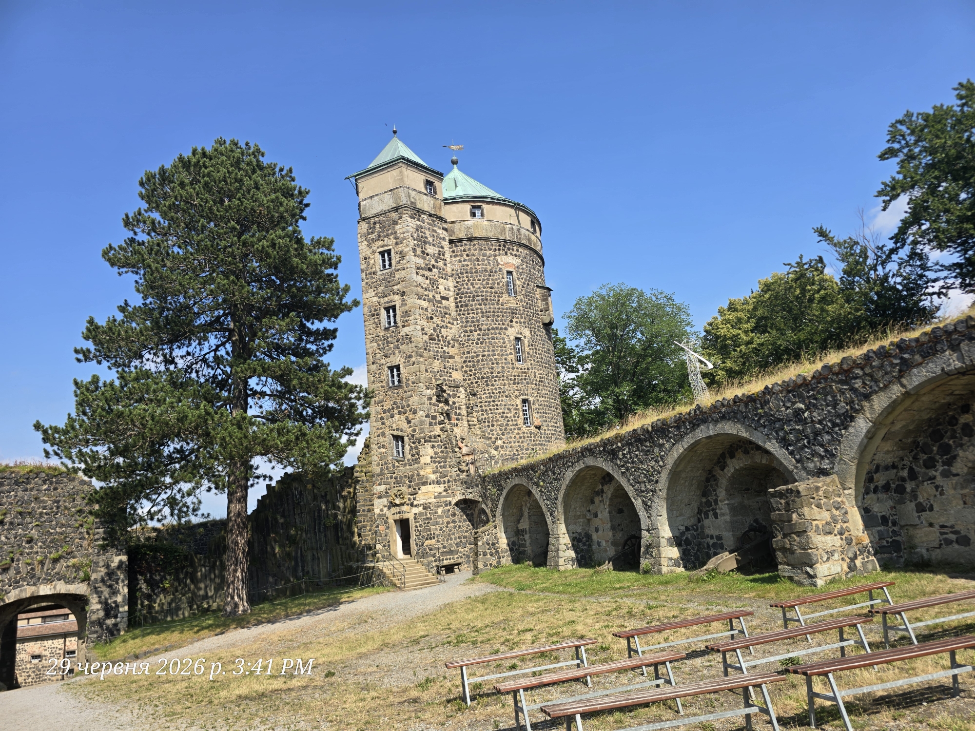 The image shows a historical stone tower with a green conical roof against a clear blue sky. The tower is surrounded by ancient stone walls, some of which are crumbling, and an arched walkway. A large evergreen tree stands to the left of the tower, and other trees and greenery are visible in the background. In the foreground, there are several rows of wooden benches, suggesting the area might be used for events or as a viewing point. The lighting indicates it is daytime, likely mid-morning or early afternoon. The scene appears tranquil and historical, evoking a sense of permanence and the passage of time. Visible text in the bottom left corner reads "29 червня 2026 р. 3:41 PM" which appears to be a date and time stamp. While no people are visible, the presence of the benches implies a past or potential gathering. The overall setting is a historic landmark, possibly a castle or fortress, in Stolpen, Germany.