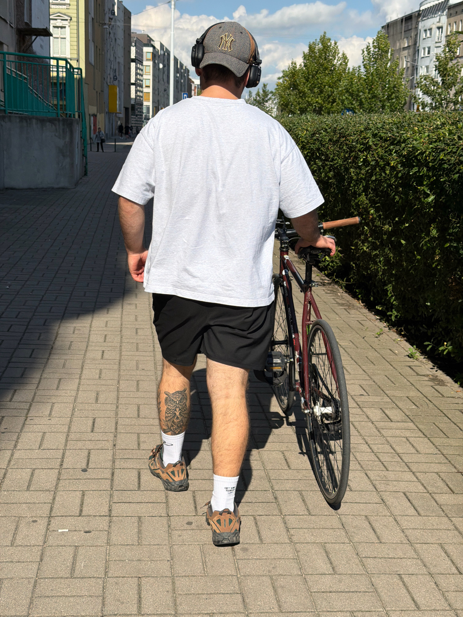 A person wearing a light gray t-shirt, black shorts, white socks, and brown athletic shoes walks on a brick-paved sidewalk, holding the handlebars of a dark red bicycle. The person has headphones on and a New York Yankees baseball cap. A tattoo of an owl is visible on their left calf. Buildings and a hedge line the sidewalk. The scene is likely in a residential area of the city of Wrocław, Poland, given the architecture and the specific design of the bicycle which appears to be a fixed-gear bike often used in urban environments. The sky is blue with scattered clouds, and the sun casts shadows on the ground.