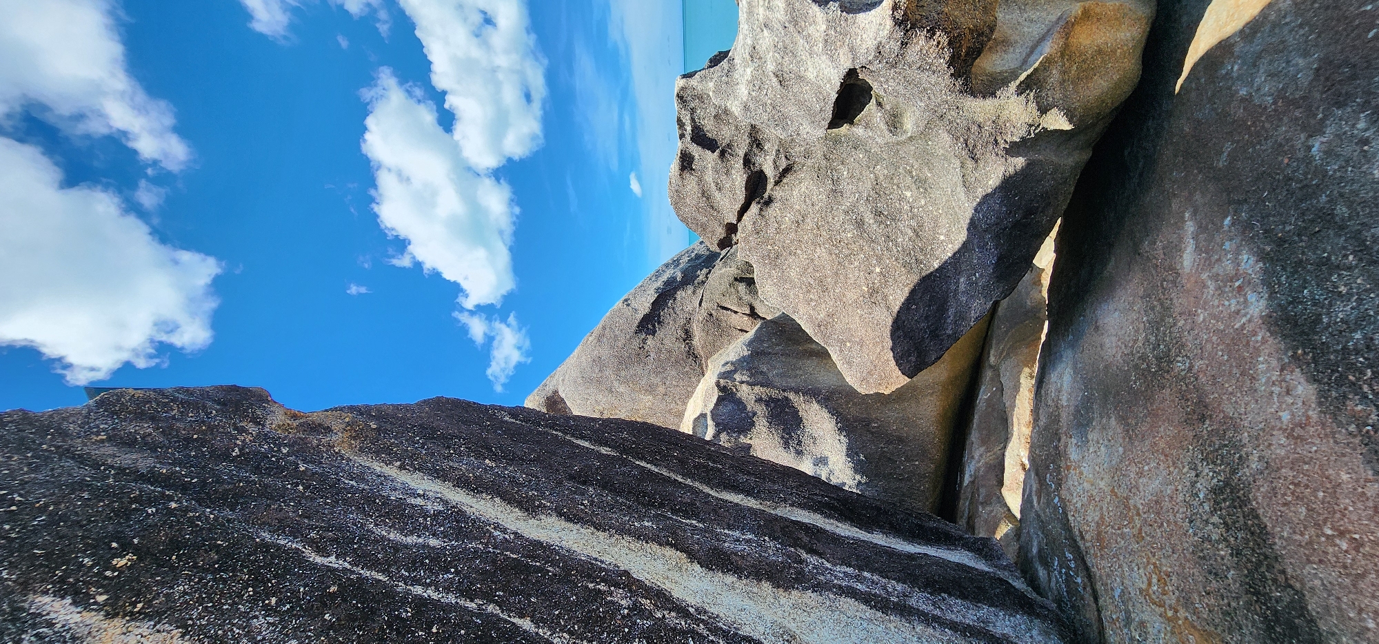 A close-up, low-angle, vertical shot captures a cluster of large, weathered granite boulders on a beach in Baan Chaweng Noi, Thailand. The foreground is dominated by the textured surface of a dark, grey boulder with vertical striations and lighter mineral deposits. To the right and behind this, other rounded, lighter grey boulders are visible, some casting shadows. Sunlight illuminates the scene, highlighting the rough surfaces of the rocks. In the upper portion of the frame, a bright blue sky is dotted with fluffy white cumulus clouds. Beyond the boulders, a sliver of turquoise ocean is visible, meeting the horizon. The scene is devoid of any people or animals, presenting a natural coastal environment.