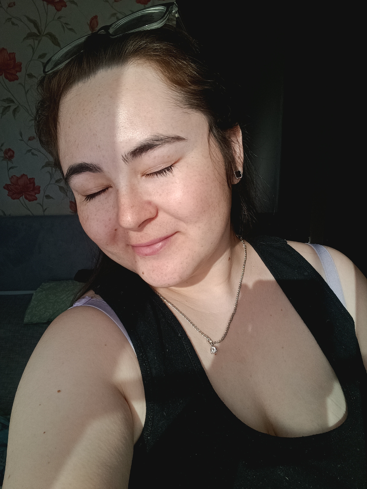 A medium shot depicts a woman with fair skin and dark hair styled back, wearing glasses perched on her head. She is wearing a black tank top over a lavender camisole and a silver necklace with a diamond pendant. The woman’s eyes are closed, and she is smiling gently. A strip of sunlight illuminates her forehead and cheek, while the rest of the image is in shadow.
The background includes a floral wallpaper, a couch, and what appears to be a pillow. The setting seems to be indoors. I do not see any indications in the image to indicate that this image was taken specifically in Noviy, Russia.