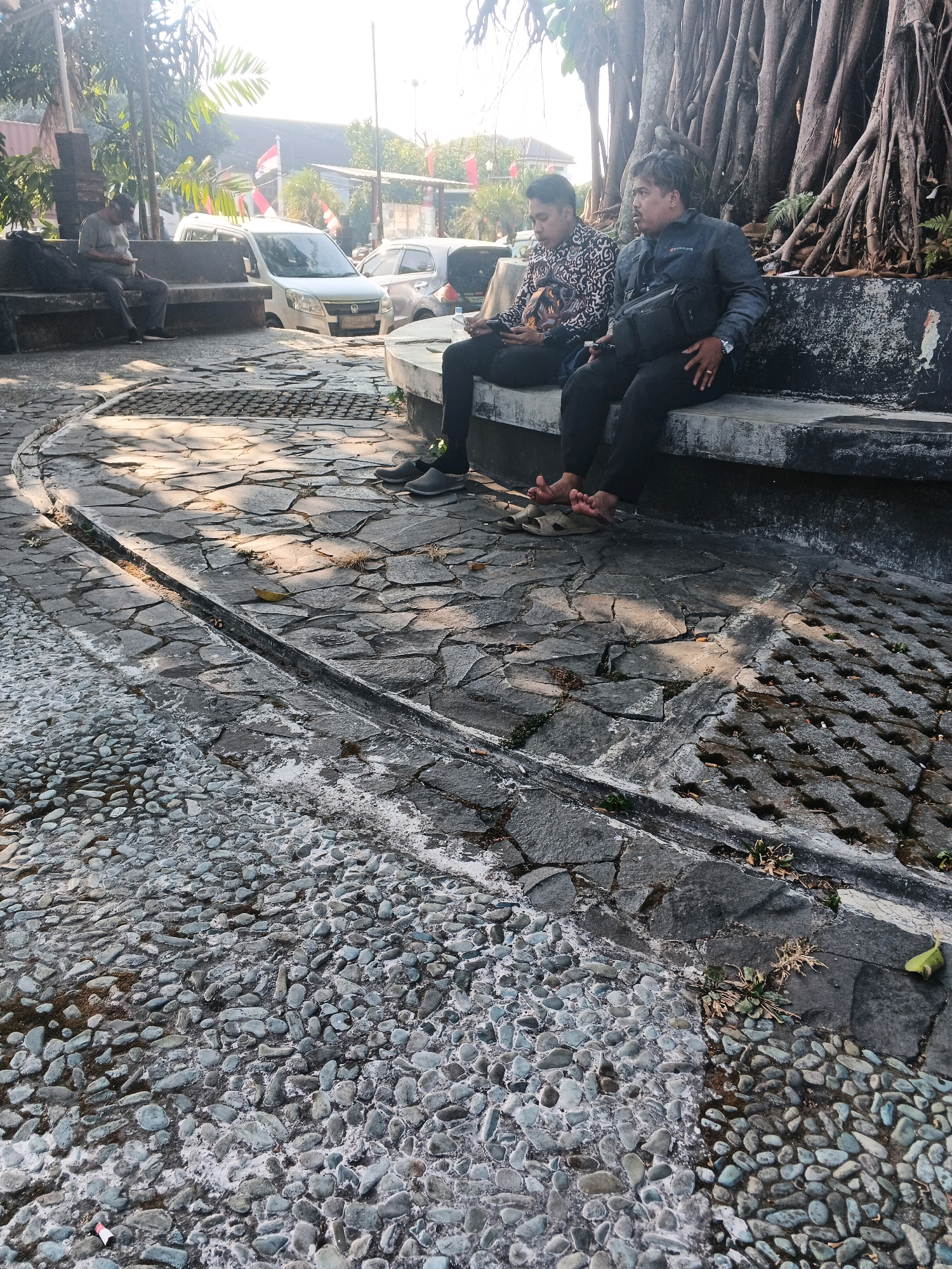The image depicts a sunny outdoor scene in Ungaran, Indonesia. Two men are seated on a concrete bench in the foreground, beneath the roots of a large tree. The man on the left is wearing a patterned shirt and holding a phone. The man on the right is wearing a dark jacket and has a cross-body bag. His legs are crossed, and he is barefoot. Behind them, a white van and a silver car are parked. The ground is paved with irregular stones and smaller pebbles. Several Indonesian flags are visible in the background.