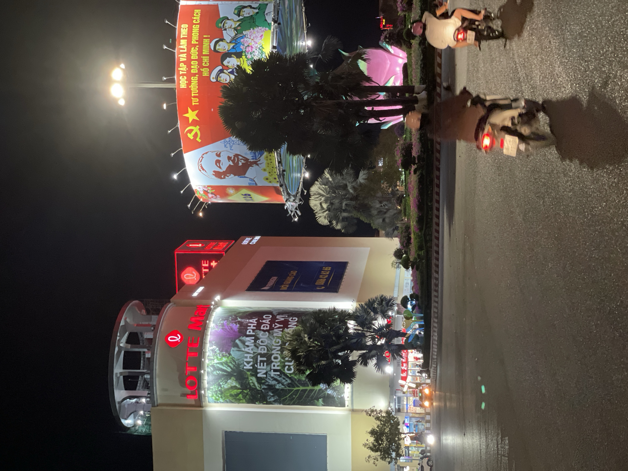 The image captures a nighttime scene in Phu Thuy Ward, Vietnam. Prominently featured is the "LOTTE Mart" shopping center with its signage illuminated in red. A large billboard displays Vietnamese communist propaganda, with images of Ho Chi Minh and slogans such as "HỌC TẬP VÀ LÀM THEO TƯ TƯỞNG, ĐẠO ĐỨC, PHONG CÁCH HỒ CHÍ MINH !" ("Study and follow Ho Chi Minh's thoughts, ethics, and style!"). Trees and decorative flora adorn the streetscape. The photograph is taken from a perspective on the side of the road, which is wet with a glistening surface. Two motorbikes are parked on the right.