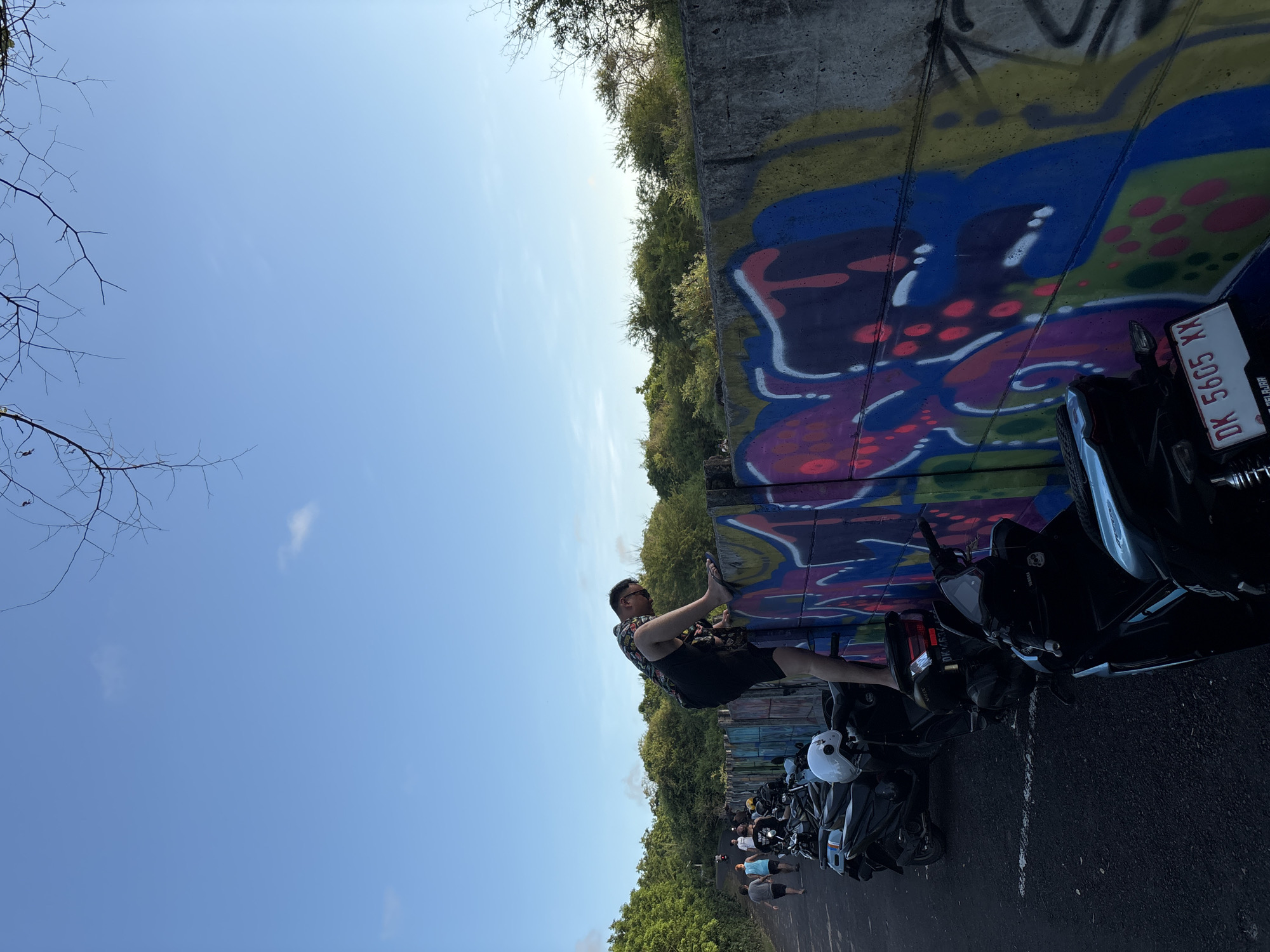A low-angle, outdoor shot shows a group of people and parked motorbikes next to a graffiti-covered concrete structure. A man stands with one leg propped against the graffiti, wearing a patterned shirt and dark shorts. The graffiti consists of vibrant blues, purples, reds, and greens. Several black motorbikes are parked on the paved surface, some with white helmets resting on them. Other people are visible in the background, some walking. The sky is clear blue with some tree branches visible in the upper-left corner. The license plate on one of the motorcycles reads "DK 5605 XX". The location is Kuta Selatan, Indonesia.