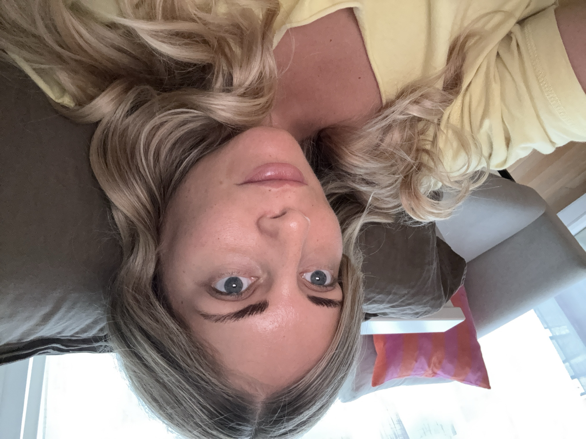 A person lies on a pillow on a sofa. The person is light skinned with blondish-brown wavy hair and blue eyes. The person is wearing a yellow shirt. A purple and orange pillow sits near the person on the sofa. A window is in the background. The photo was taken indoors. The scene appears to be a domestic setting. There are no indications or details that suggest the location is in Paris, France.