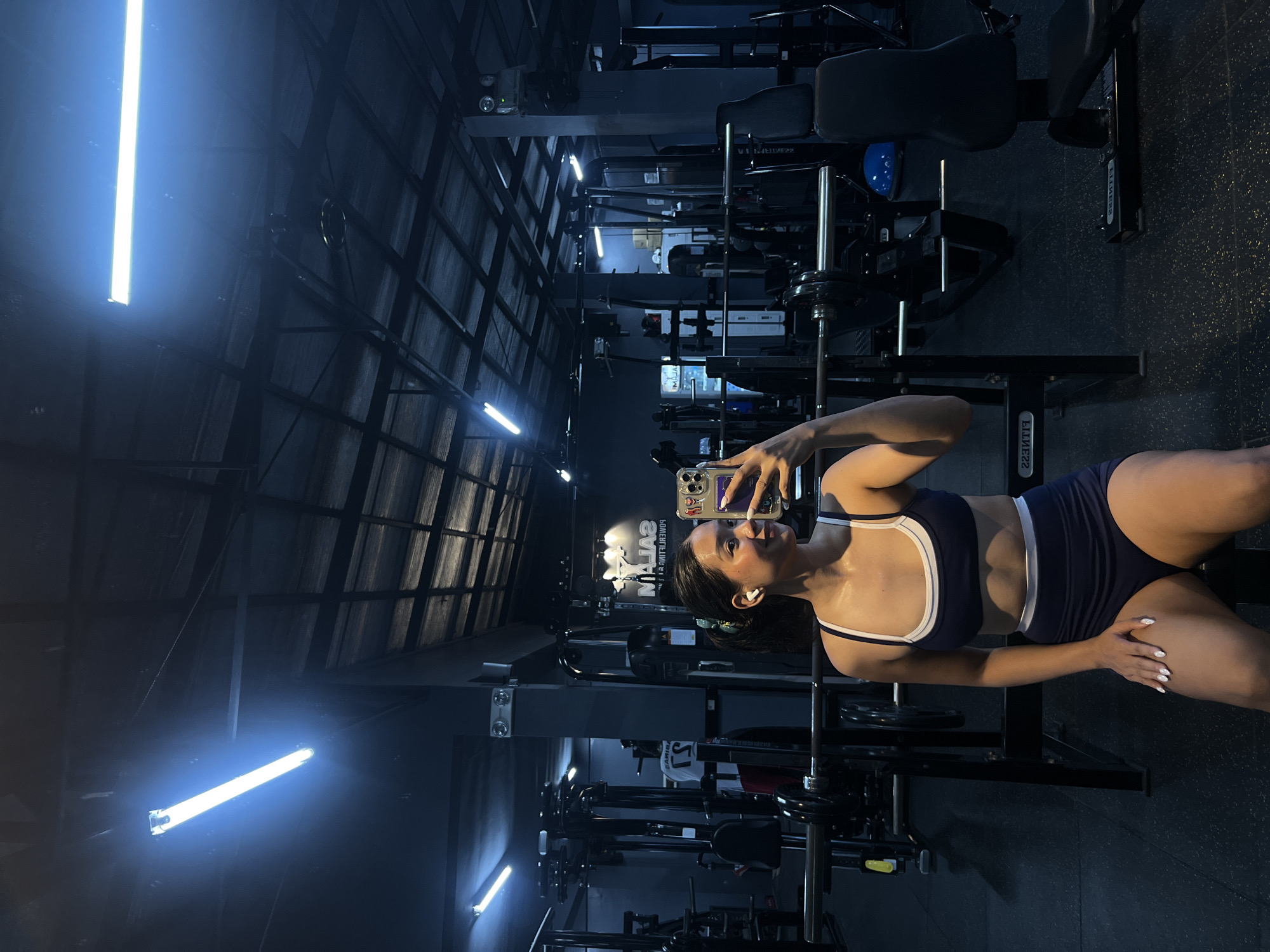 The image shows a young woman taking a mirror selfie in a dimly lit gym. She is wearing a navy blue sports bra and shorts with white trim. She is sitting on a weight bench, with barbells and weight plates visible behind her. The gym appears to be equipped with various weight machines and equipment. The overall atmosphere is dark, with fluorescent lights overhead casting a blueish hue. The gym is located in Tanza, Philippines.