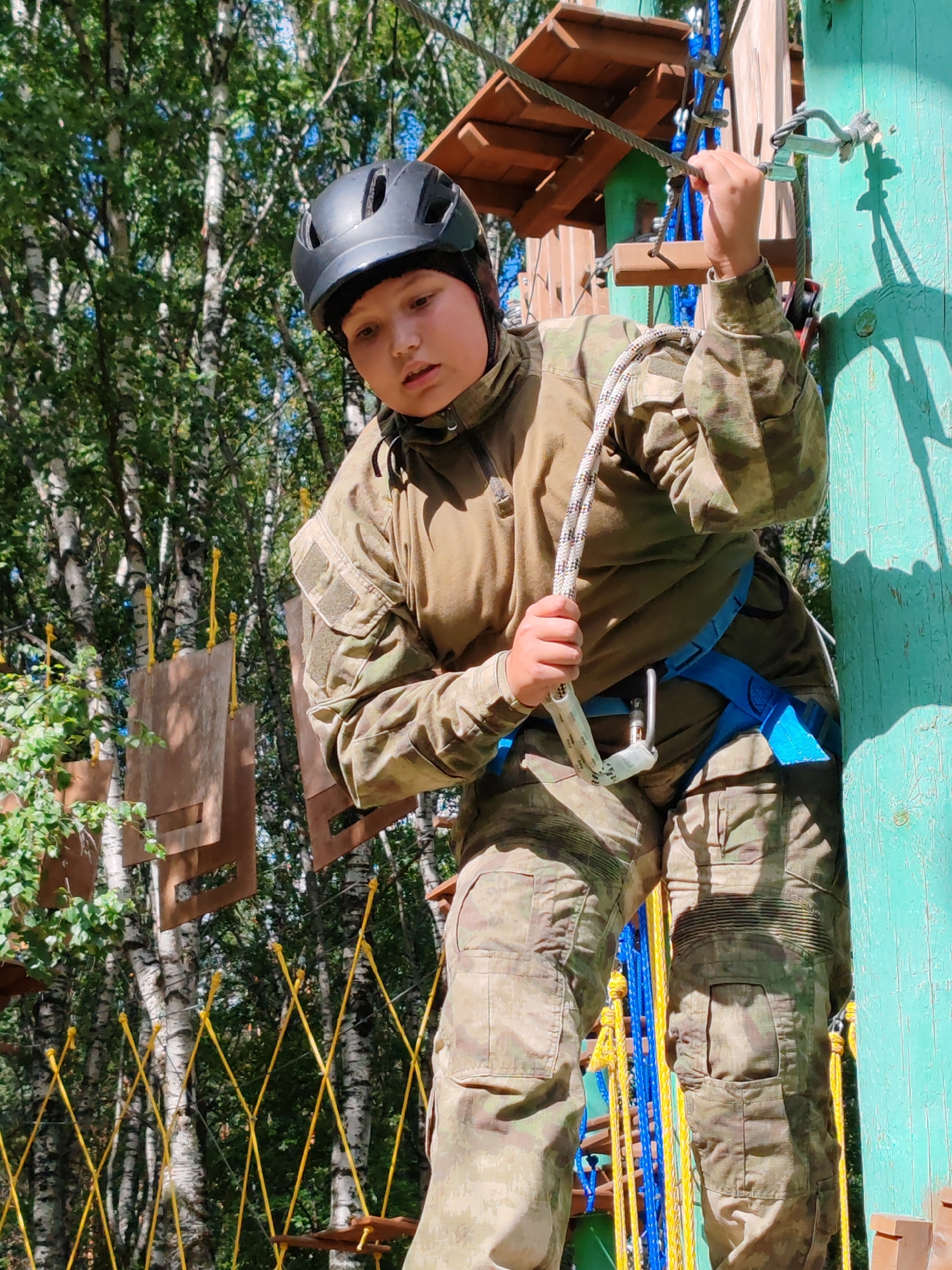 A young person wearing a black helmet and camouflage clothing is navigating an obstacle course in a forest. They are secured by a blue harness and a rope with a carabiner, which is attached to a metal cable overhead. The course appears to be made of wood and rope elements, suspended between trees. The person is looking down, concentrating on their next move. In the background, birch trees with green foliage are visible under a bright sky. The scene is set in the Komi Republic National Park, located in the city of Syktyvkar, Russia.
