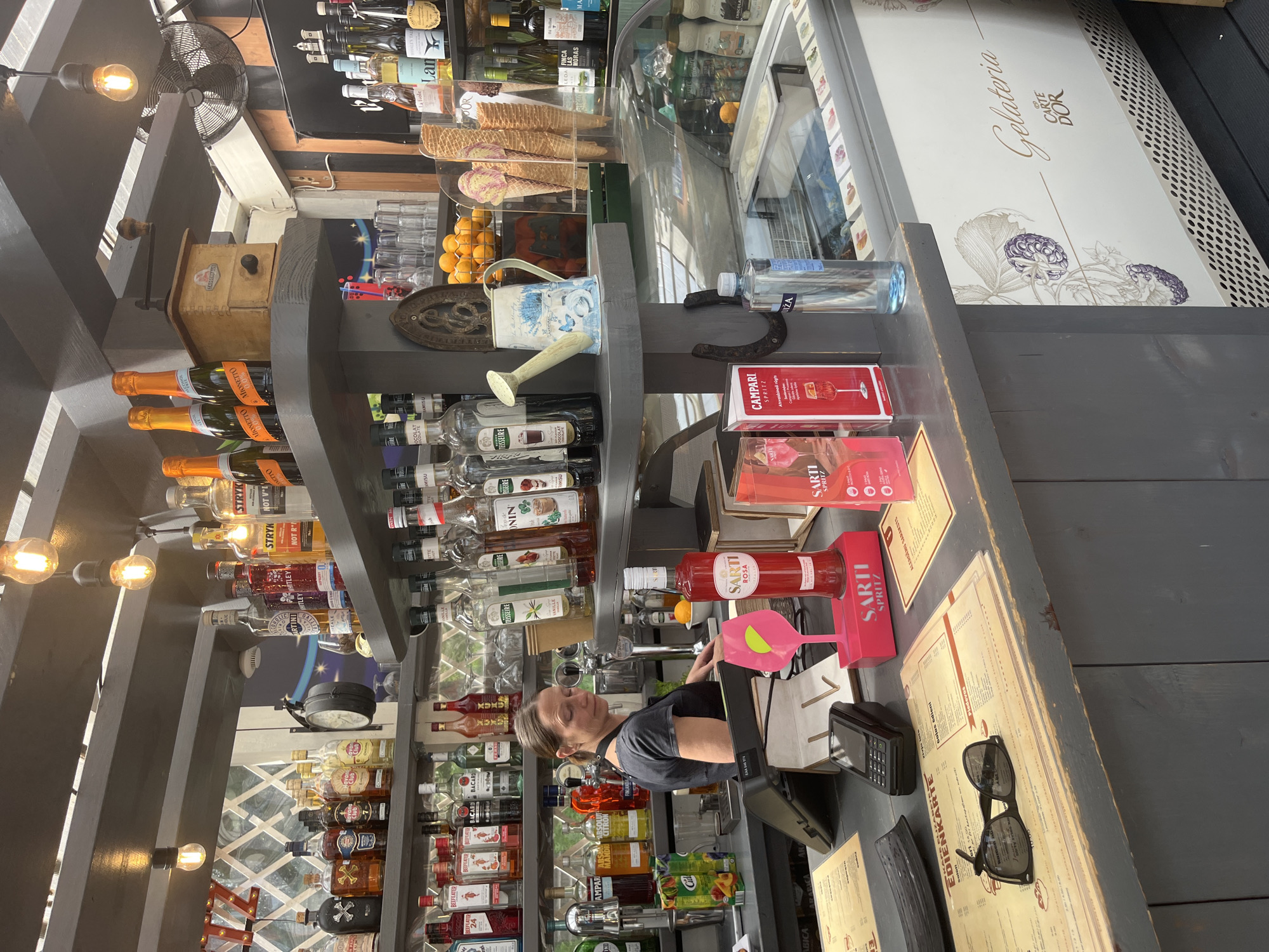 In Riga, Latvia, a woman with brown hair, wearing a dark grey apron over a dark t-shirt, is working behind a bar in an outdoor establishment. The bar is stocked with various bottles of alcohol, including what appear to be liqueurs, wines, and spirits, arranged on shelves. There are also bottles of sparkling wine and other beverages displayed prominently. In front of the woman, on the counter, are a credit card reader, a pink cocktail glass with a green garnish, and several boxes and menus. The establishment has an ice cream display case with tubs of ice cream and cones. In the background, there are more shelves filled with bottles and various decorative items, including a vintage coffee grinder and a watering can. The lighting suggests it's daytime, with warm light bulbs hanging from the ceiling. The overall atmosphere appears to be that of a casual bar or cafe.