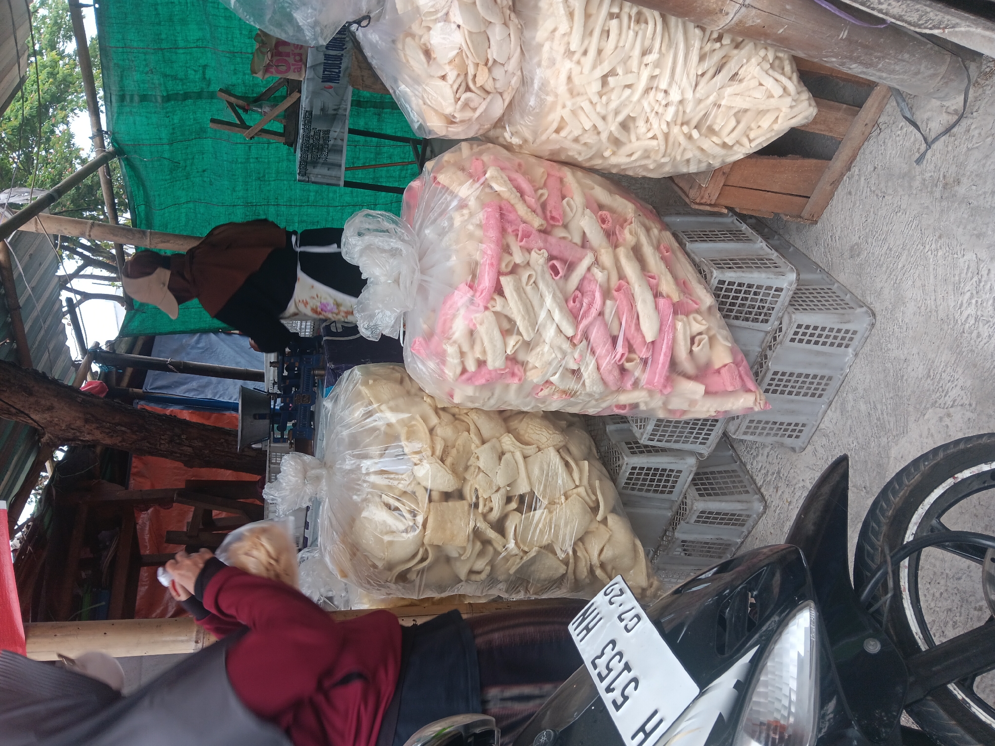 A street vendor in Batursari, Indonesia, is shown selling bagged snacks. In the foreground, a motorcycle is parked. Behind it, several large plastic bags are filled with puffed snacks, some are white and square, others are pink and tubular. A person wearing a hijab and a maroon jacket is holding a bag of snacks, appearing to be serving a customer. Another person, wearing a brown jacket and a cap, is visible in the background. The scene is outdoors, under a makeshift shelter with a green tarpaulin. Various baskets and wooden crates are used to display the snacks. The ground is concrete. The overall setting suggests a typical roadside market.