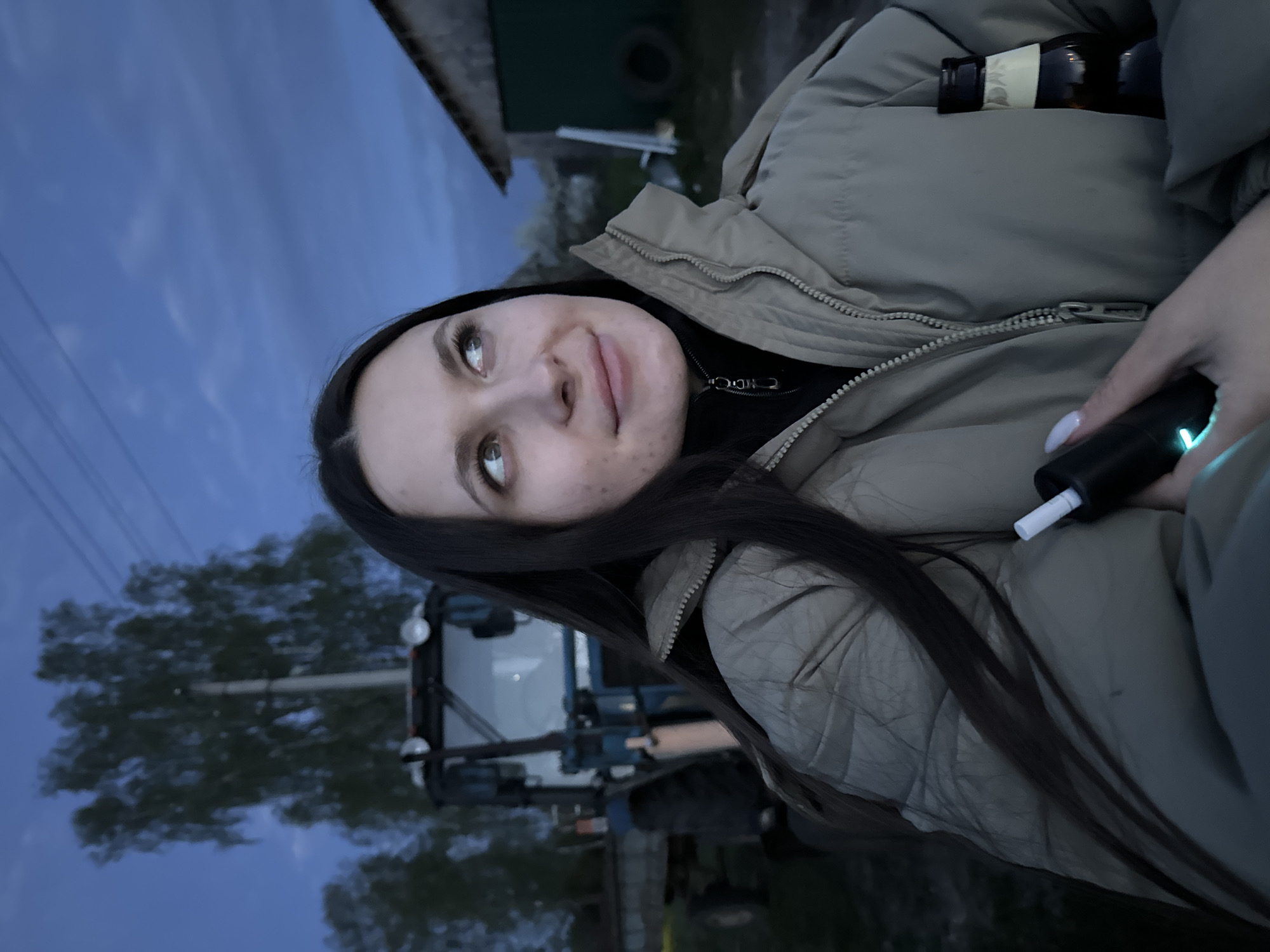 A young woman with long, dark hair is depicted outdoors at dusk in Красный Маяк, Russia. She is wearing a light brown puffer jacket and a black turtleneck sweater. Her gaze is directed upwards and to the left. She is holding a black device with a white cigarette protruding from the top in her right hand, and a bottle is visible in the right pocket of her jacket. A blue tractor is partially visible in the background, along with trees and buildings. The sky is dark blue with some clouds.