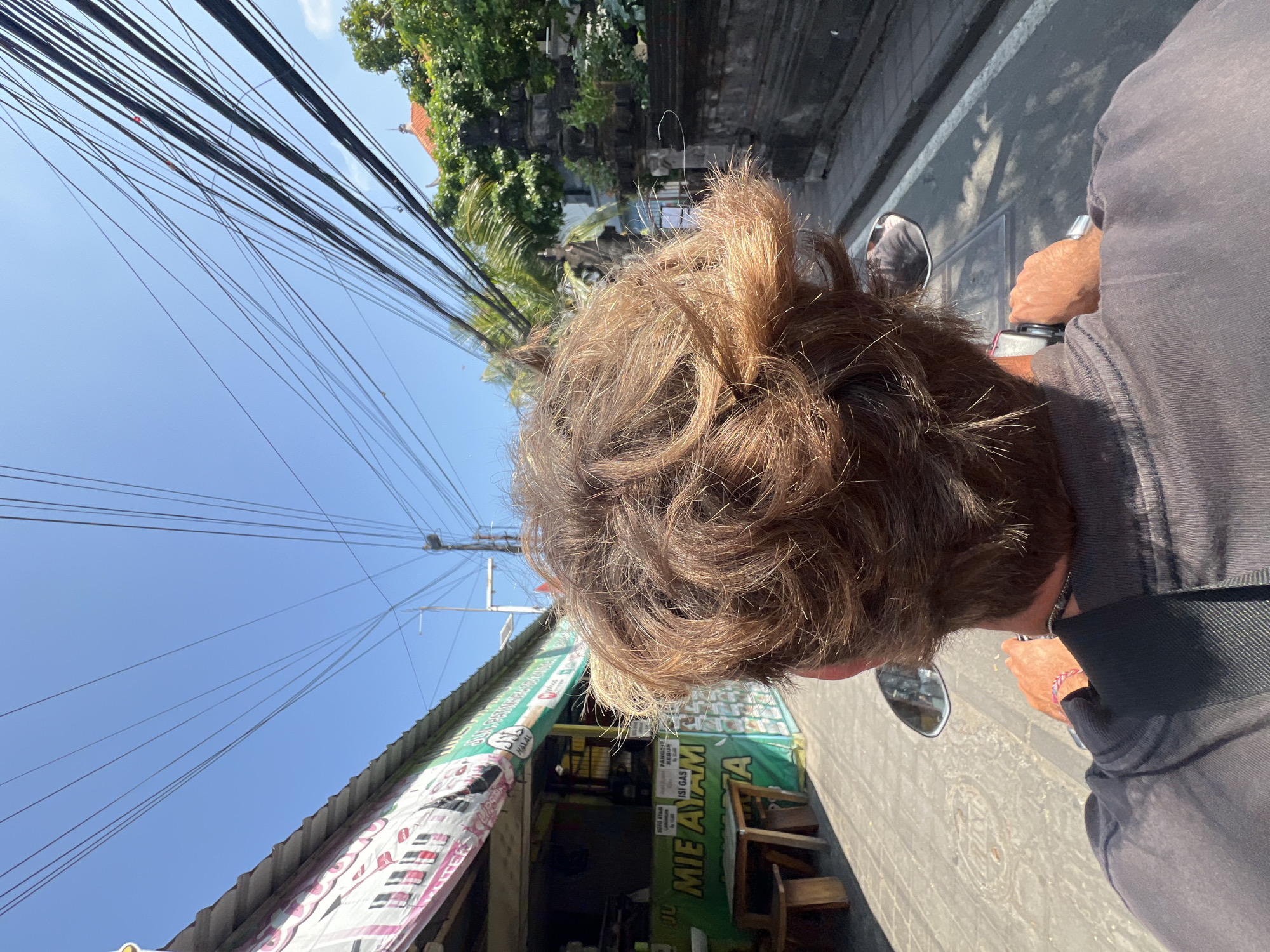 An over-the-shoulder, first-person perspective shot shows a person with light brown, tousled hair riding a motorbike through Kuta, Indonesia. The rider's head is in the foreground, with a black shirt visible below. They are holding the handlebars, and their right hand is in focus. The road is paved with grey and white markings. On the left side of the road, a food stall with a green awning is visible. A sign in Indonesian reads "MIE AYAM JAKARTA". Various menus and posters are displayed on the stall. To the right, a stone wall and lush green trees can be seen. Above, a bright blue sky is filled with a dense network of power lines.