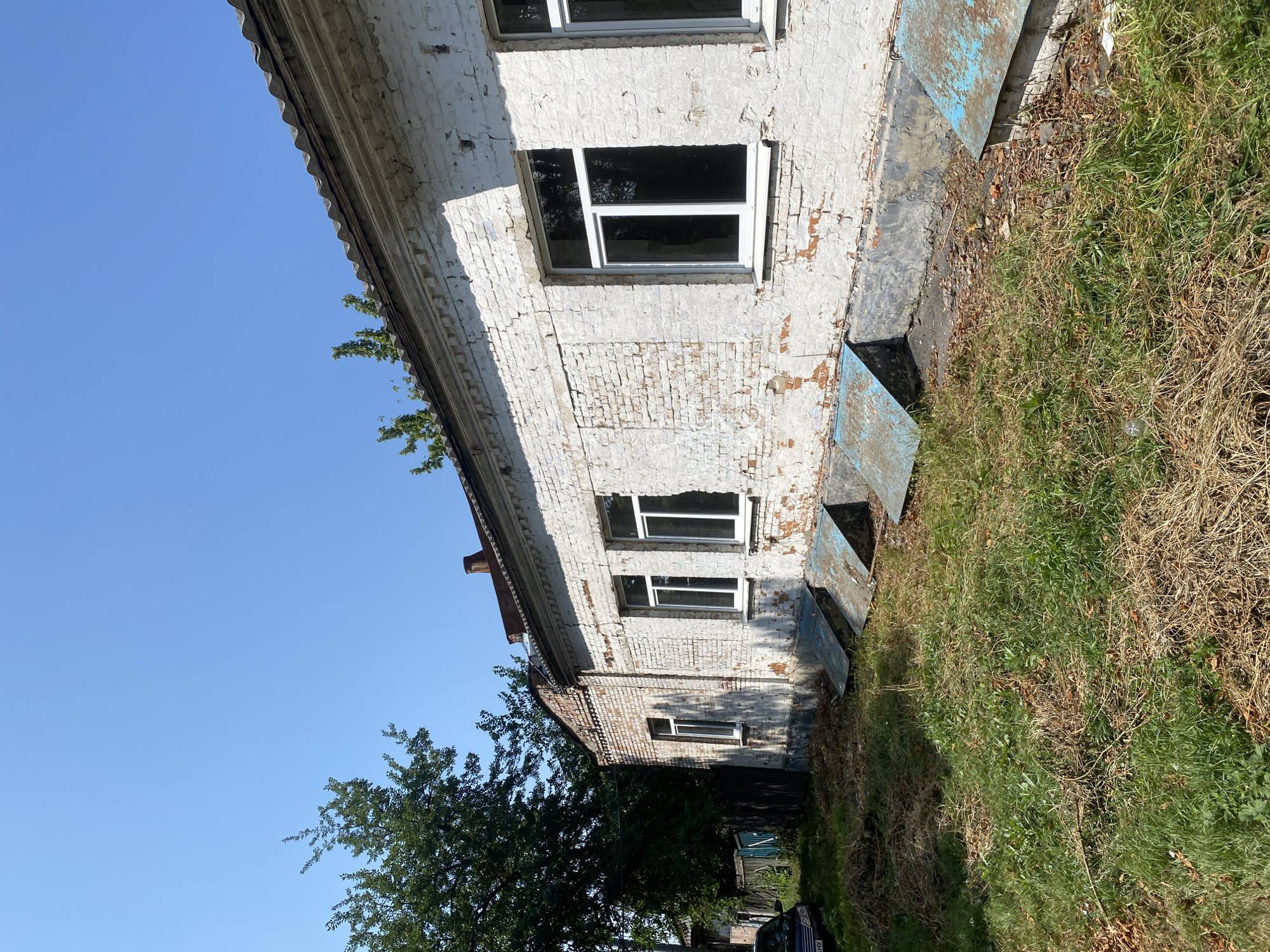 This is a photograph of an old, partially dilapidated brick building with white-painted walls and a corrugated roof. The building has several windows with modern white frames, some of which appear to have been recently installed. In front of the building, there are several basement access hatches with rusty blue metal covers. The ground is covered in overgrown grass, dry foliage, and debris. To the left of the building, there is a large, dense tree and a section of a wooden fence. A dark-colored car is partially visible on the left edge of the frame. The sky is clear and blue. The scene appears to be in a rural or semi-urban area, likely a neglected part of the city of Poltava, Ukraine, given the context of the request. There are no people visible in the image.