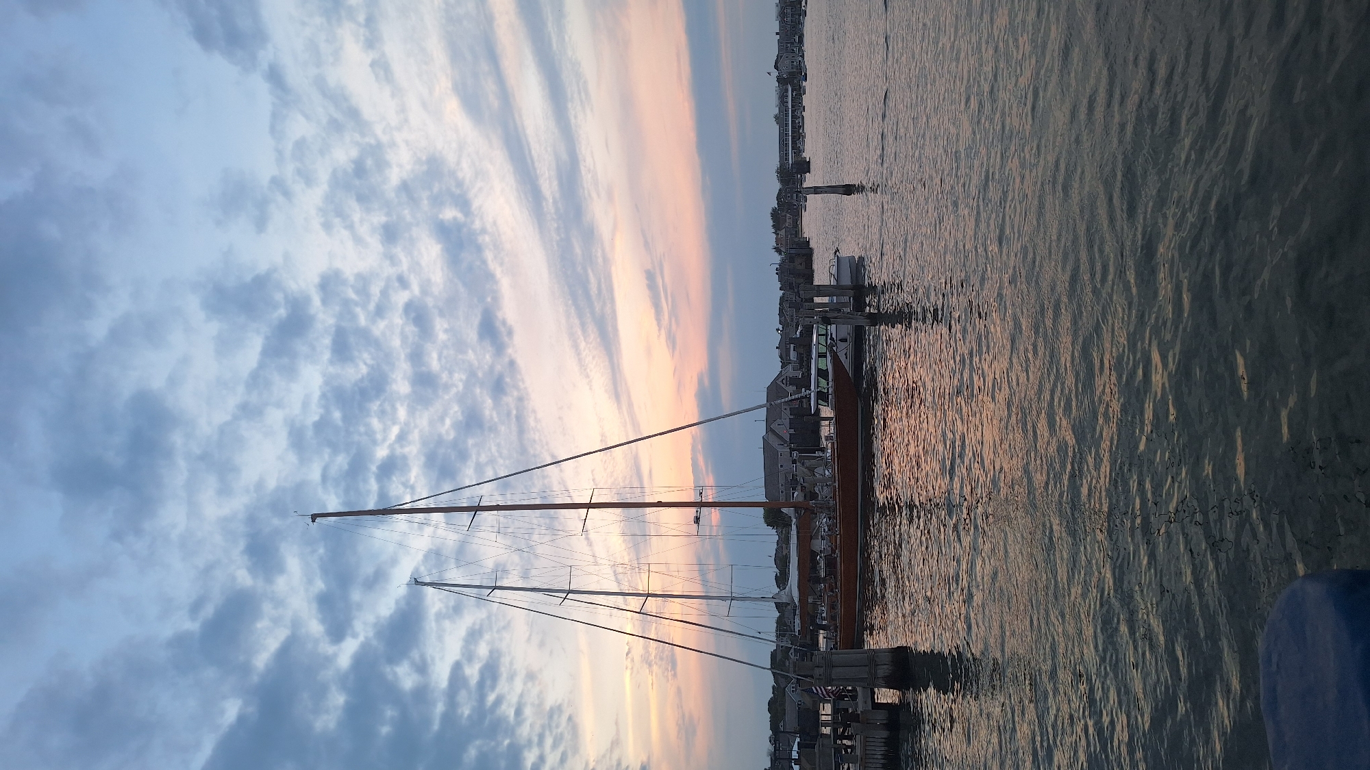 An exterior, eye-level, vertical medium shot captures a harbor scene at dusk on Nantucket, United States of America. A large, wooden sailboat with two masts is docked to the left, its reflection visible in the calm water. Another smaller white boat is docked to the right of the sailboat. In the background, houses line the shore. The sky is filled with scattered clouds, with hues of pink and orange from the setting sun visible near the horizon. The water is a deep blue-gray, with gentle ripples. No people are visible in the image.
