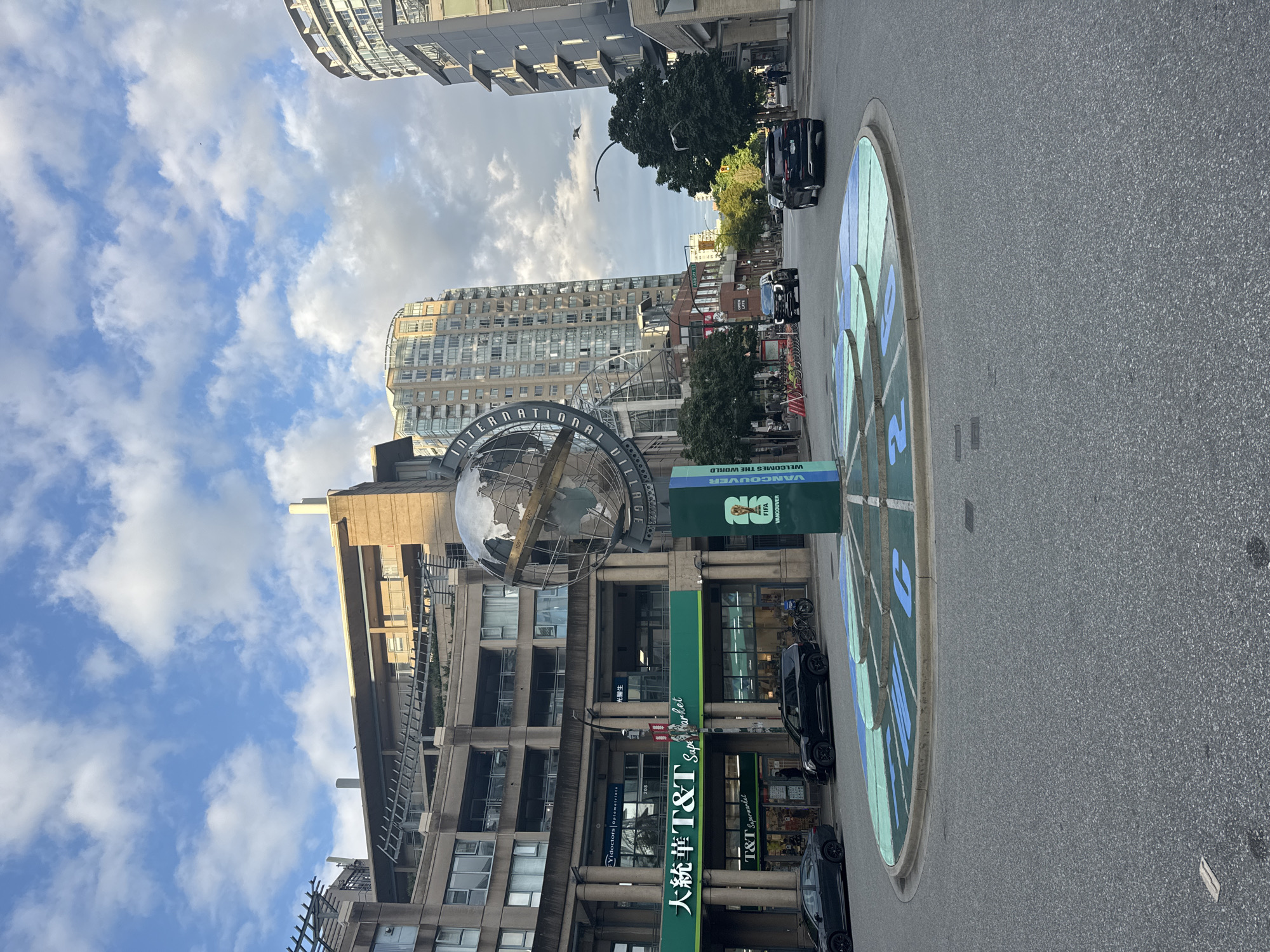 This is a full, eye-level, outdoor, street-level shot taken in Vancouver, Canada, at a roundabout featuring a prominent globe sculpture. The globe is a metallic sphere, depicted as a world map, mounted on a central pillar with signage. Surrounding the globe is a circular plaza with segmented sections painted in green and blue, featuring large blue numbers and what appear to be stylized clock markings.
In the foreground, a grey asphalt roundabout is visible, with the edge of the painted plaza in the mid-ground. Two black SUVs are driving on the road. The left SUV is in the foreground, and the right SUV is further down the road, heading away from the viewer. On the left side of the image, a large T&T Supermarket storefront is visible, with green and white signage. Next to it are entrances for "Eye Doctors Optometrists" and another establishment with Chinese and English signage. Behind the storefronts, a multi-story building with a curved facade and balconies is visible.
The background is dominated by tall, modern buildings, some with curved architectural features, and interspersed with trees. The sky above is a mix of blue with scattered white clouds, suggesting daylight. A street lamp with a curved arm is visible extending over the road. The overall scene depicts an urban environment in a city setting.