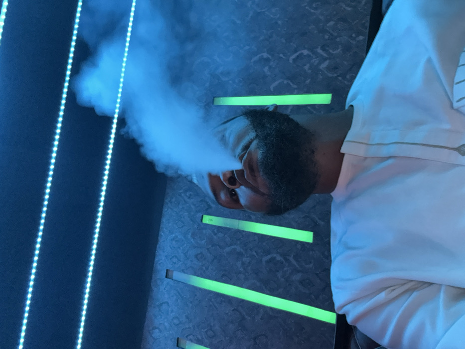 A bearded man, adorned in a white shirt, is depicted exhaling a dense cloud of vapor. His face is illuminated by a blue-green light. The background is a textured wall, punctuated by horizontal bars of light green. To the side is a vertical line of closely spaced blue lights. The image is slightly tilted counter-clockwise.