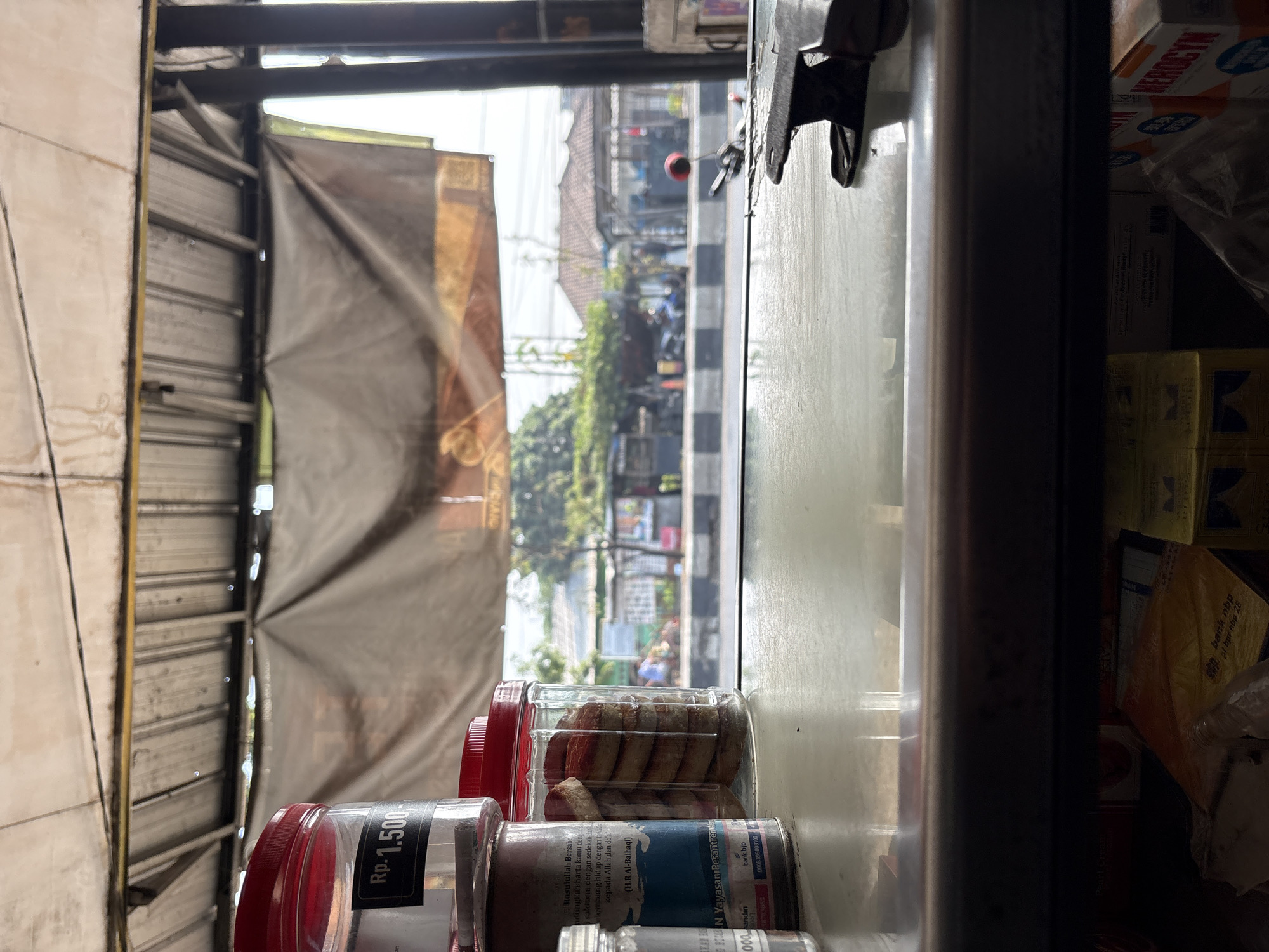 The image captures a street-level view from an indoor vendor stall in Palimanan, Indonesia. In the foreground, a reflective surface, likely a counter or table, occupies the lower portion of the frame. To the left, stacked containers are visible; one clear plastic jar with a red lid contains stacked cookies, and a label on the jar mentions "Yayasan Pesantren". Another clear container to its left has a black label displaying "Rp. 1.500-". On the right side of the counter, a dark metal clip is positioned.
Beyond the counter, the scene opens to an outdoor environment. A corrugated metal roof structure extends overhead, supported by dark vertical posts. A beige tarp hangs behind the stall, obscuring some of the background. The view through the opening reveals a street with a black and white striped pedestrian crossing. Several motorcycles and a few people are present on the street. Buildings with tiled roofs are visible in the middle ground, surrounded by trees and greenery. The overall impression is that of a busy roadside stall in a tropical urban or semi-urban setting.
