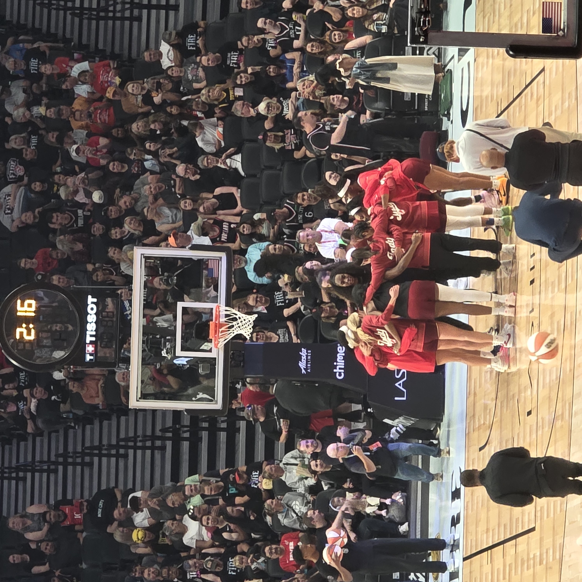 A basketball team in red uniforms huddles on a polished wooden court in a stadium filled with spectators. The players have their arms around each other, and some are wearing headbands. A basketball rests on the court in the foreground. The spectators are seated in tiered bleachers behind a black railing. Some are holding cell phones and cameras. A large digital clock above the court displays "2:16". The scoreboard also shows "TISSOT". The overall scene suggests a professional basketball game is in progress or has recently concluded. This scene is likely taking place in Portland, Oregon, United States, given the presence of the word "PORTLAND" on some of the spectators' attire.