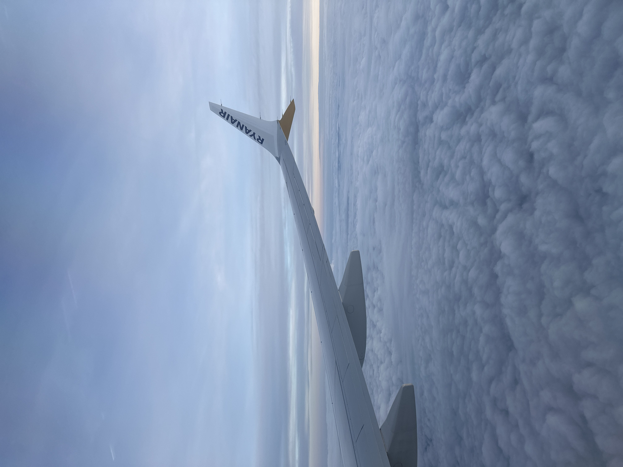 The image presents an aerial view from an airplane window, showcasing the aircraft's wing extending across the frame against a backdrop of clouds and sky. The main subject is the white airplane wing, which features a prominent upward-curving winglet at its tip. On the inner surface of this winglet, the word "RYANAIR" is clearly visible in dark blue capital letters, identifying the airline. The very tip of the winglet is accented with a small section of yellow or gold. Further along the wing, two engine nacelles are partially visible beneath it.
The scene depicts the plane flying high above a dense, textured layer of white and grey clouds, which cover the lower half of the view and extend to the horizon. Above this cloud layer, the sky transitions from a light, hazy blue near the clouds to a clearer, deeper blue at the top. A subtle band of warmer light, suggesting either dawn or dusk, is visible where the sky meets the distant cloud horizon.
The activity taking place is air travel. Notable details include the peaceful and expansive atmosphere created by the high-altitude perspective. The time of day appears to be either early morning or late afternoon due to the soft, diffused light and the warm hues on the distant horizon. There are no specific location cues beyond the airline branding. The image is presented with a vertical orientation, as if the camera was held sideways, but captures a typical horizontal view from an aircraft.