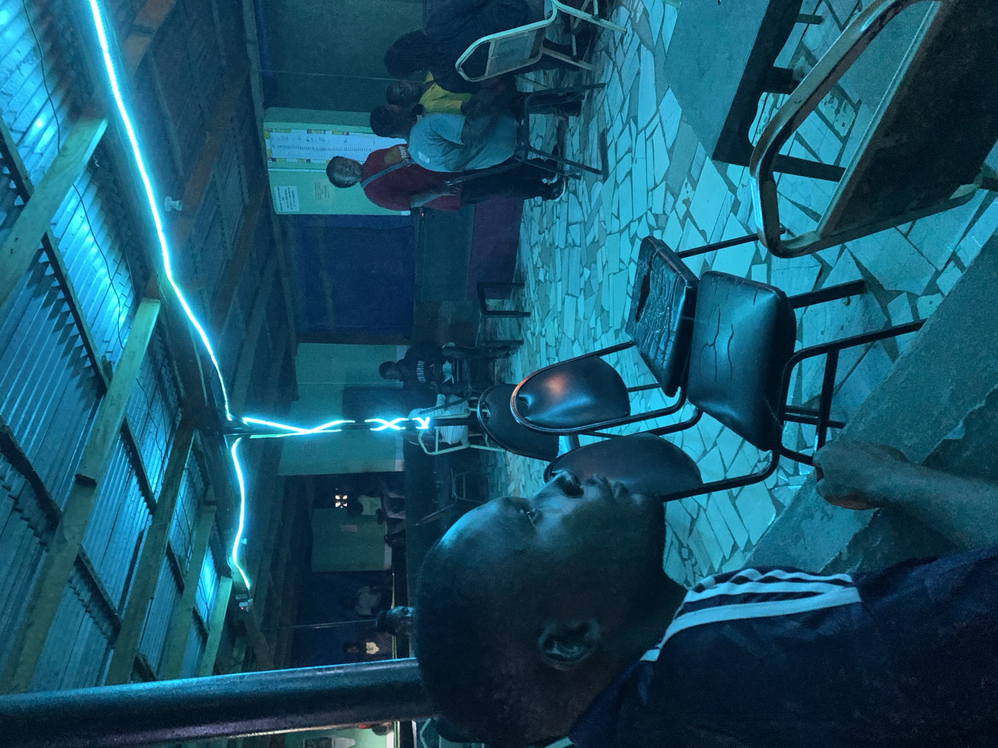The scene depicts the interior of a building in the vicinity of New Stadium, Nigeria. Several people are sitting on chairs arranged in rows. The foreground features a person in a dark blue shirt with white stripes, with their face and upper body visible. The building has a corrugated metal roof illuminated by a blue light fixture. The floor is tiled with a mosaic pattern.