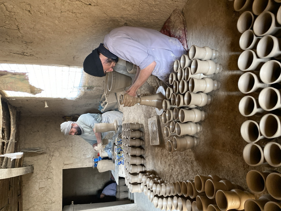 This image depicts the interior of a pottery workshop in Kandahar, Afghanistan. Two adult males are actively engaged in pottery creation.
In the foreground-right, a man with a dark beanie, dark scarf, and beard, wearing a light-colored long-sleeved shirt, sits on a red cushion on the packed dirt floor. He operates a pottery wheel with his hands, shaping a tall, light-brown clay vessel. To his left, a second man with a white turban and beard, wearing a light-colored shirt and a white apron, is similarly seated and working at another pottery wheel where a finished or nearly finished vessel stands. He appears to be preparing or refining clay. In the dimly lit background-left, visible through an open doorway, a third, indistinct individual is seated on the floor.
The workshop features rough, unpainted mud-brick walls, a packed dirt floor, and an exposed wooden beam ceiling with hanging fabric. Natural light enters through a gridded window on the right side of the main room and the open doorway. Hundreds of newly fashioned, unfired light-brown clay vessels, primarily cups and vases of varying sizes and designs, are systematically arranged in multiple rows across the dirt floor, in various stages of drying. A small collection of finished, decorated ceramic bowls with blue, white, and yellow patterns is visible on a low platform near the turbaned potter. The scene portrays a functional, traditional pottery manufacturing environment.
