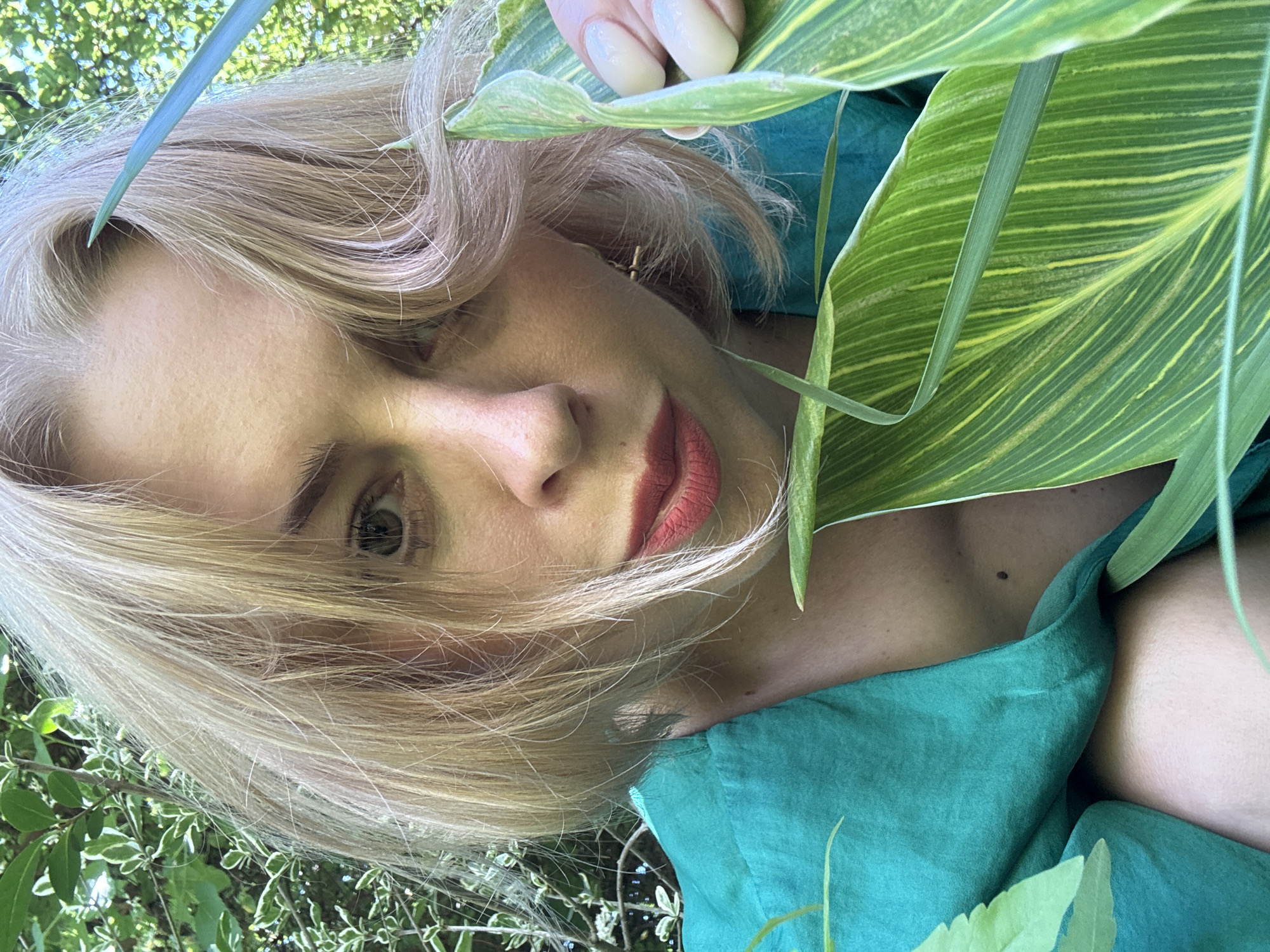 The image shows a close-up of a woman with blonde hair and red lipstick, partially obscured by large green leaves. She is wearing a teal garment. The background suggests an outdoor environment with foliage, likely a garden. The scene is brightly lit, with sunlight filtering through the leaves. There is no visual information to suggest the location is in Sochi, Russia.