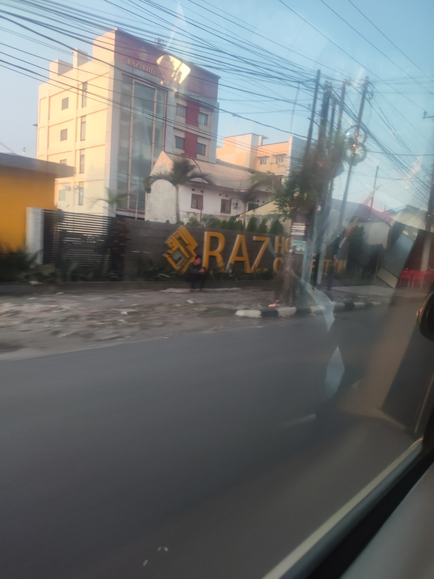 The image depicts the exterior of RAZ HOTEL in Medan, Indonesia, as seen from a moving vehicle. A multi-story building with a modern facade, featuring a combination of cream-colored panels and reflective glass, is the primary subject. The hotel name "RAZ HOTEL" is prominently displayed in large, yellow, three-dimensional letters against a dark wall. In front of the hotel, a row of palm trees and green foliage lines the property. A paved road runs parallel to the hotel, with a sidewalk and some rubble visible between the road and the hotel's entrance. A person wearing a dark jacket and pants is seen squatting near the large yellow letters, possibly resting or observing. Electrical wires and utility poles are visible in the background, suggesting an urban environment. The sky is a clear, light blue, indicating daytime. The overall scene captures a typical street view in a bustling city, with the hotel serving as a landmark.