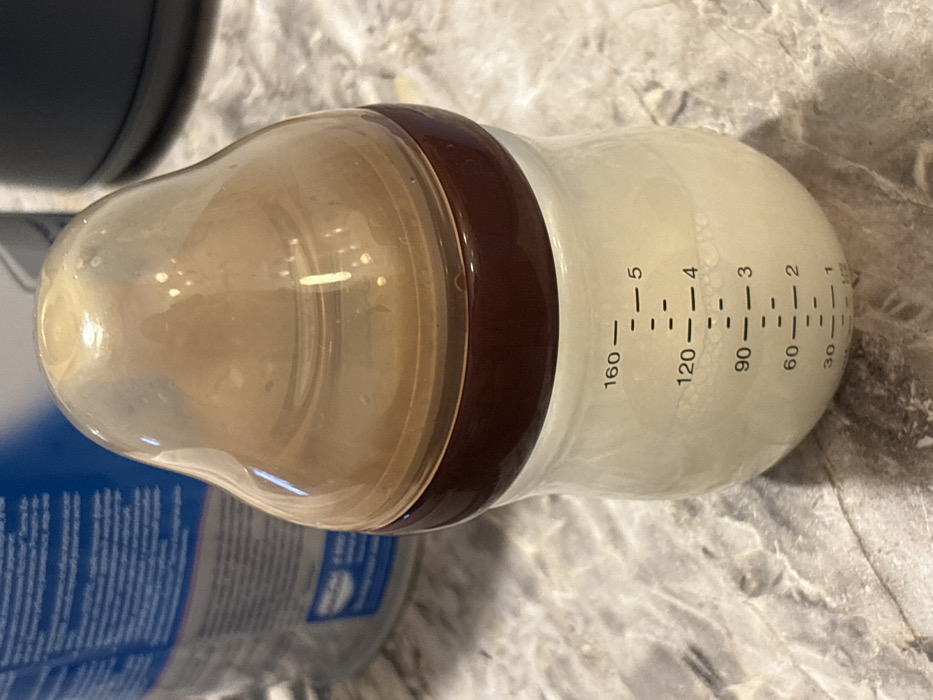An upward-angled, close-up photograph displays a transparent baby bottle, filled with an opaque white liquid (likely formula or milk) to a level between the 120 and 160 milliliter markings. The bottle features a dark brown neck ring and a translucent, yellowish-brown, bell-shaped nipple. Visible volume measurements include 30, 60, 90, 120, and 160, with corresponding marks 1 through 5. Small bubbles are present within the liquid. The bottle rests on a light-colored, mottled countertop, suggestive of granite or marble. In the blurred background, a blue and white container, possibly a can of infant formula, is partially visible to the left. A dark, cylindrical object is partially visible to the right. The scene is static and appears to be indoors in Lezhnevo, Russia.