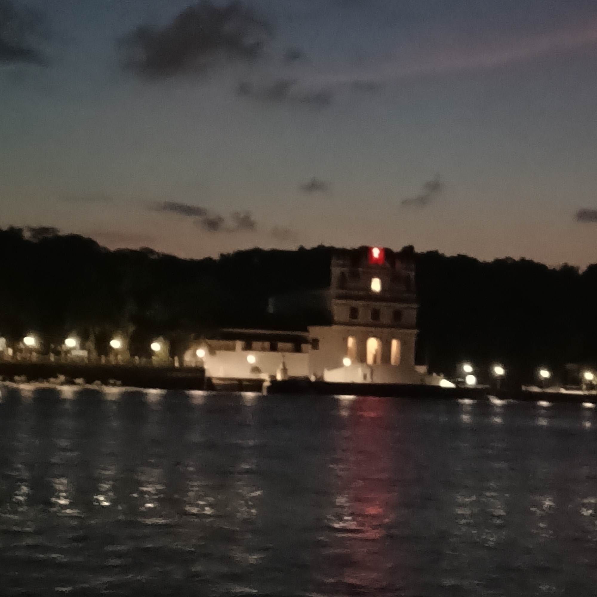The image shows the Reis Magos Fort at night, located in Britona, Goa, India, as viewed from the Mandovi River. The fort is illuminated with lights from within, including a red light on the roof. The surrounding environment is dark, with trees visible along the shoreline and lights lining the waterfront. The river reflects the lights from the fort and shoreline. There are no people visible. The sky is a mix of dark blues and oranges, suggesting either sunset or twilight.