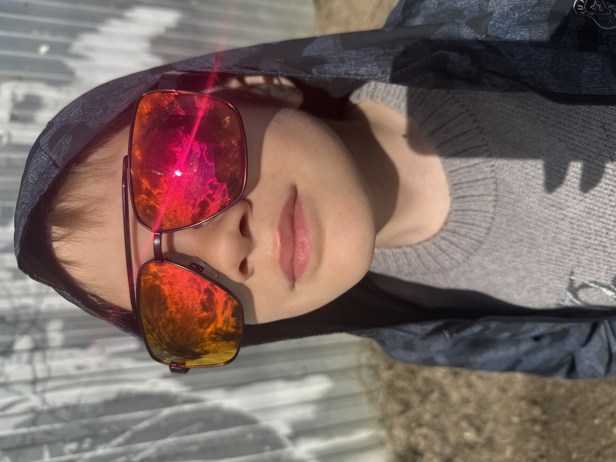 A close-up shot shows a person wearing a gray sweater and a dark hooded jacket. The person is also wearing sunglasses with red and orange lenses. The background appears to be a corrugated metal surface and the ground with some dry vegetation. It is not possible to determine if this image was taken in Dmitrov, Russia.