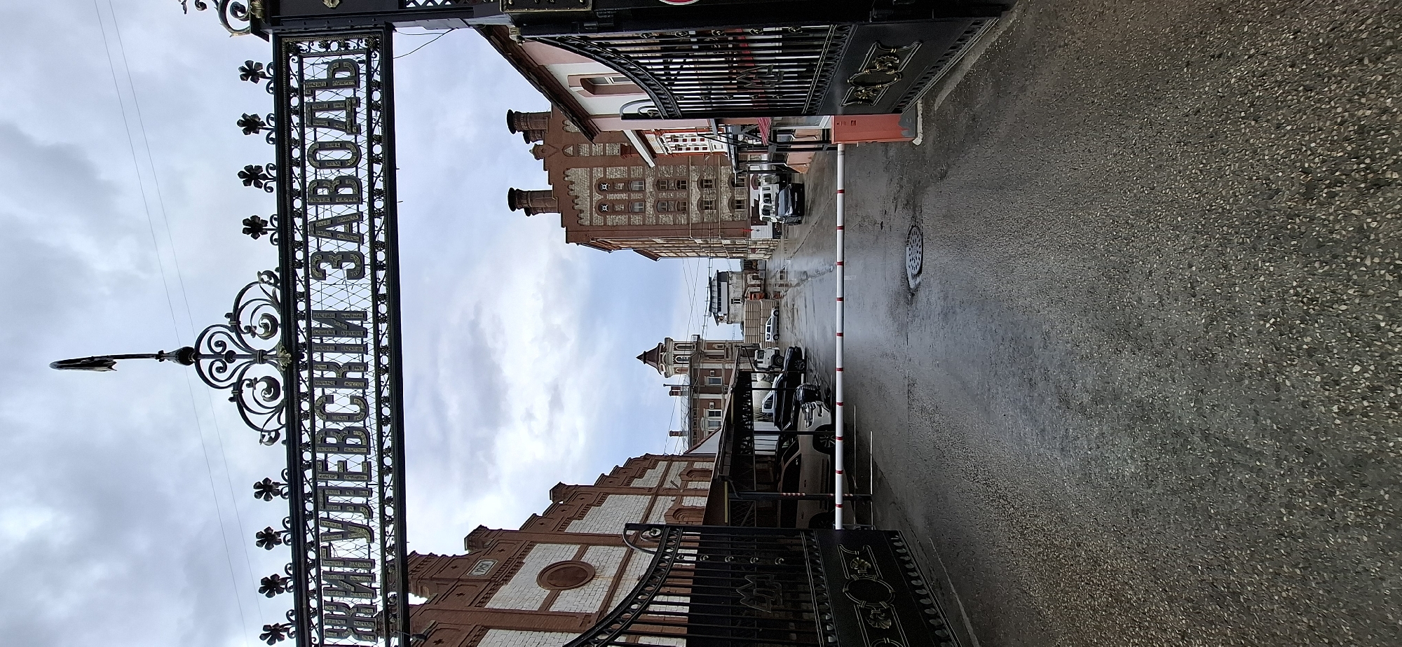 The image depicts the entrance to the Zhigulevskiy Zavod (Zhigulevsky Factory) in Samara, Russia. The foreground is dominated by a wet asphalt road leading into a courtyard. A red and white striped barrier gate is lowered across the road, blocking access. On the left and right sides of the entrance, ornate black metal gates stand open, with decorative elements.
The architecture of the factory buildings is visible beyond the gates. The building on the left features red brickwork with white accents and visible windows. A sign on this building indicates "1901". The building on the right is also constructed with red brick and has several windows. In the background, a tower with a distinctive tiered roof stands out, along with other factory structures and chimneys. Several vehicles, including cars and a truck, are parked under a covered area on the left, and more vehicles are visible further down the courtyard. The sky is overcast, suggesting a cloudy day. The overall scene is that of an industrial facility entrance on a wet day.