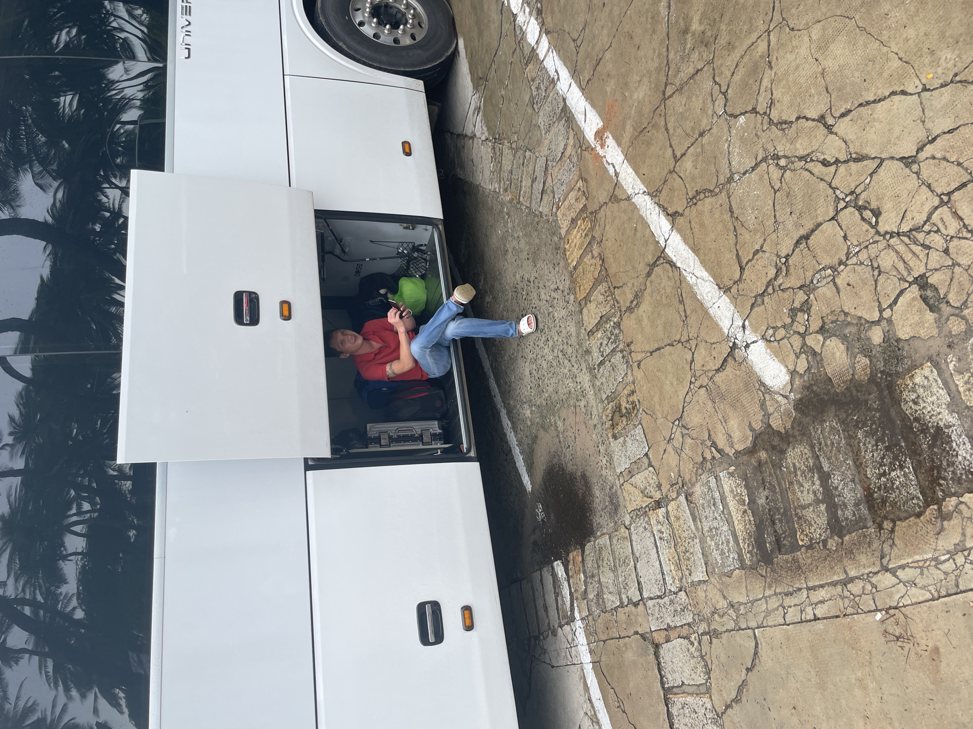 The image shows a man reclining in the open baggage compartment of a white bus parked in what appears to be a parking area. The bus has "UNIVER" visible on its side. The man is wearing a red shirt, blue jeans, and sandals. He appears to be using a mobile phone. Various luggage and equipment are visible inside the bus compartment. The ground is a cracked concrete surface with white parking lines and a section that is possibly paved with stone bricks. Phu Thuy Ward, Vietnam, is not verifiable from the image.