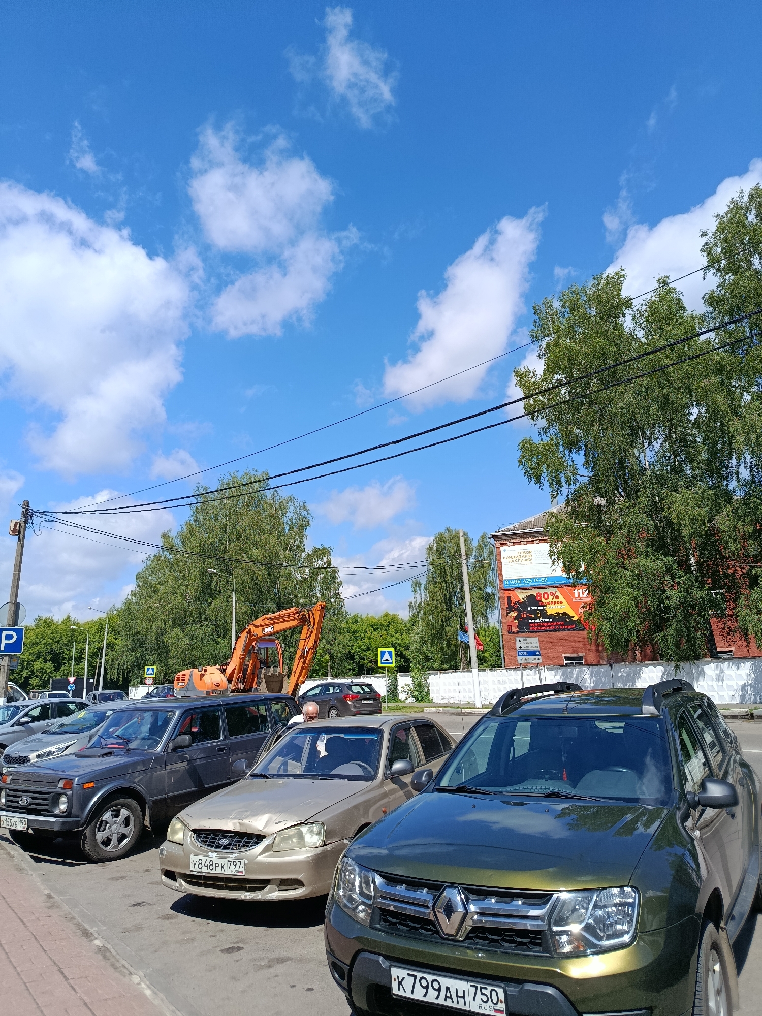 In Orekhovo-Zuyevo, Russia, on a bright, sunny day with scattered clouds, a street scene unfolds. Cars are parked along a sidewalk, with a darker colored SUV in the foreground. Behind it, a gold sedan shows visible damage to its front end. Further back, a dark gray SUV is parked. An excavator is positioned on the side of the road, its orange arm extended. In the background, a red brick building displays advertisements, and dense green trees line the street, with power lines crisscrossing overhead. A pedestrian crossing sign is visible near the excavator.