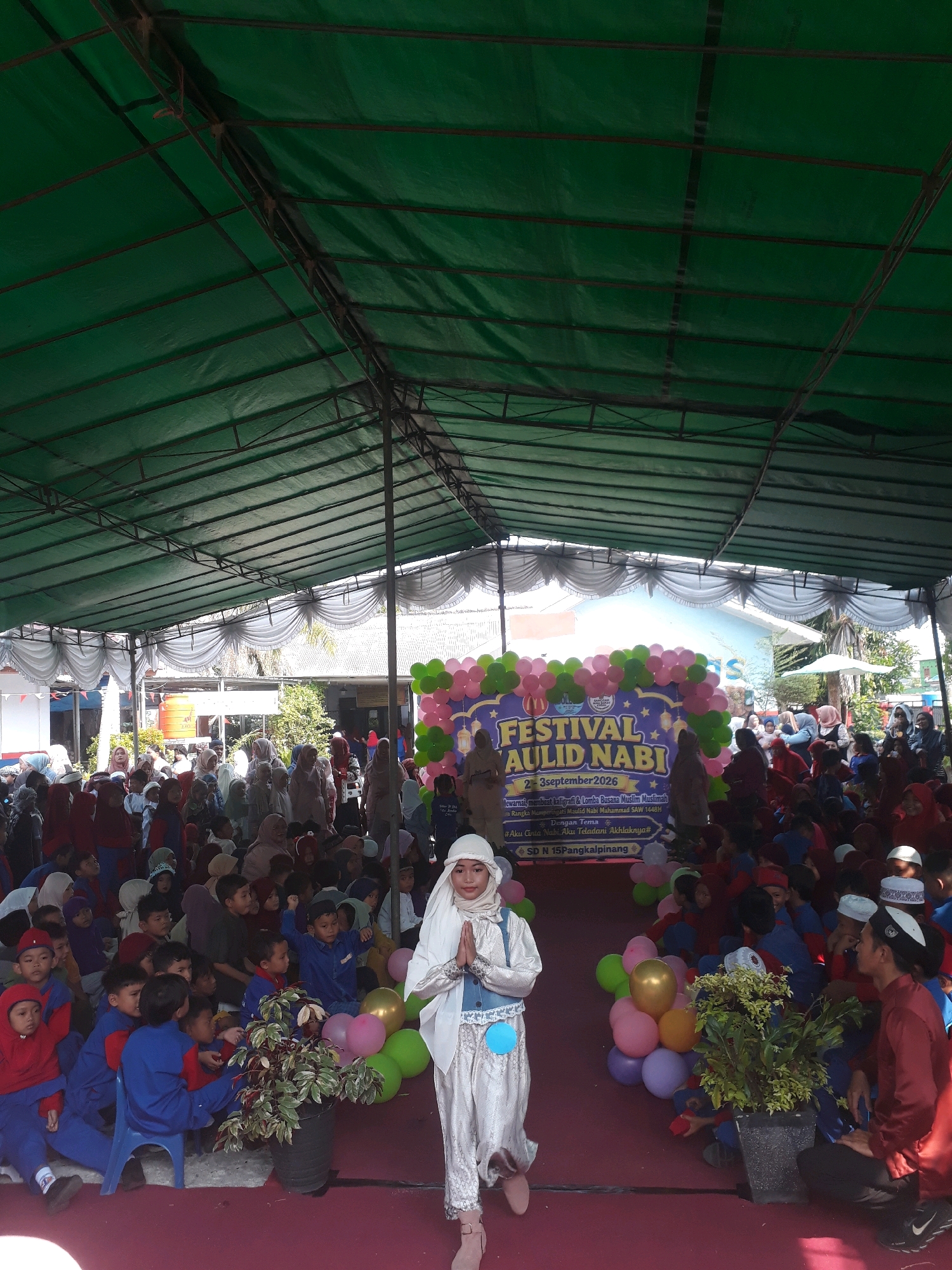 An outdoor event is taking place under a large green tarp supported by metal poles. A crowd of children and adults are gathered on a maroon-colored surface. In the foreground, a young girl wearing a white hijab and a light-colored, ornate outfit stands with her hands clasped in a prayer-like gesture. She is facing left and looking towards the viewer. To her right, a potted plant with green and red leaves sits on the ground. Behind the plant, a cluster of pink, green, and purple balloons is arranged.
A banner in the background reads "FESTIVAL AULID.NABI" with "2-3 September 2026" below it, and text in Indonesian detailing events and the theme "#Aku Ceria Nabi Aku Teladani Akhlaknya#". The banner also indicates "SD N 15 Pangkalpinang", suggesting the event is taking place at or is related to this school in Pangkalpinang, Indonesia. Many of the children are wearing uniforms, some in red and blue, and others in various shades of brown and white. The overall atmosphere appears to be festive and community-oriented.