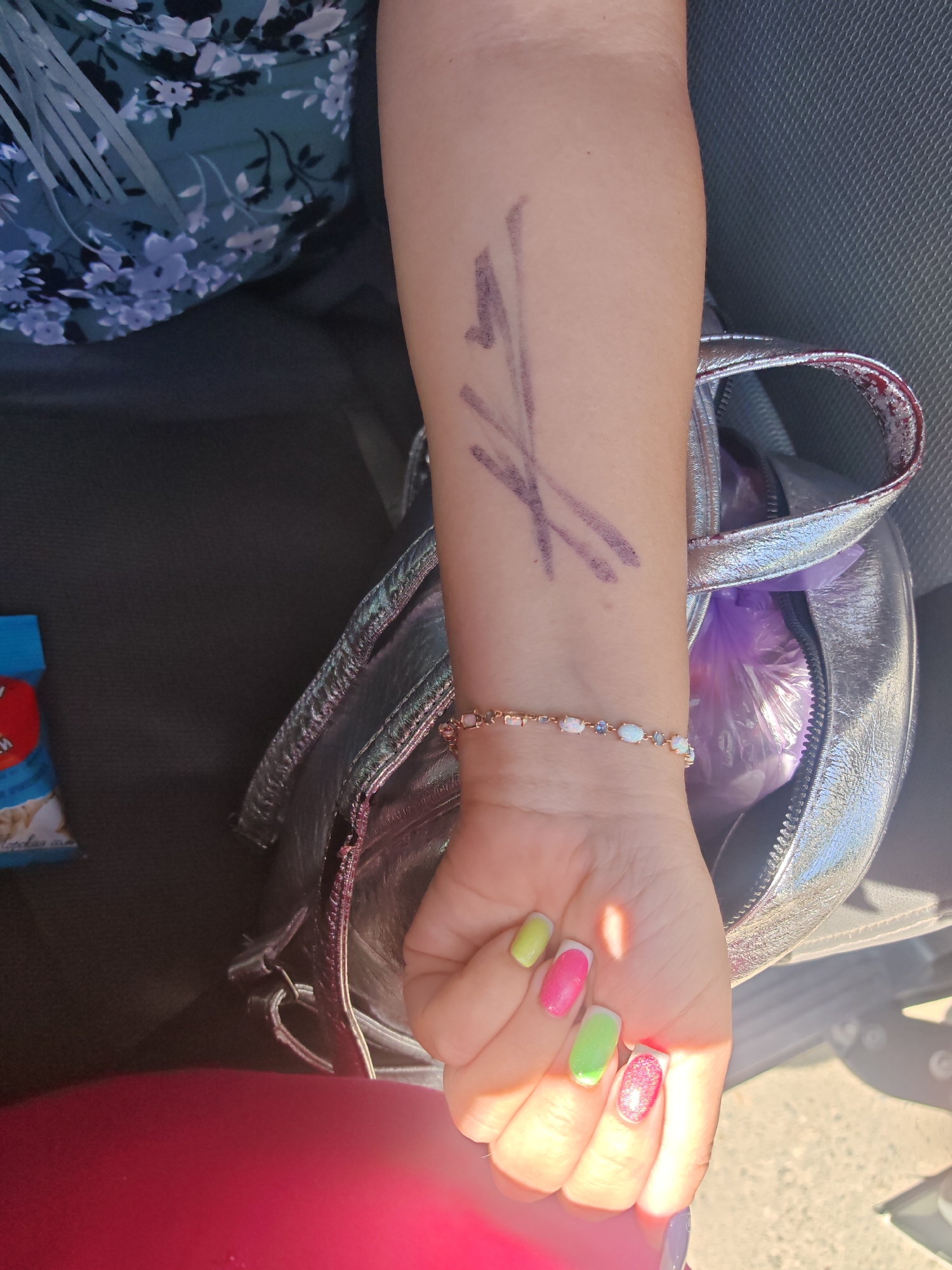 The image shows a person's arm with a purple drawing of what appears to be a stylized flower or star. The person is wearing a floral-patterned top and a silver purse. The person's nails are painted in bright colors, including pink, green, and white. The image is taken outdoors, with sunlight casting shadows. There are no visible landmarks or context that would indicate the location in Varvarovka, Russia.