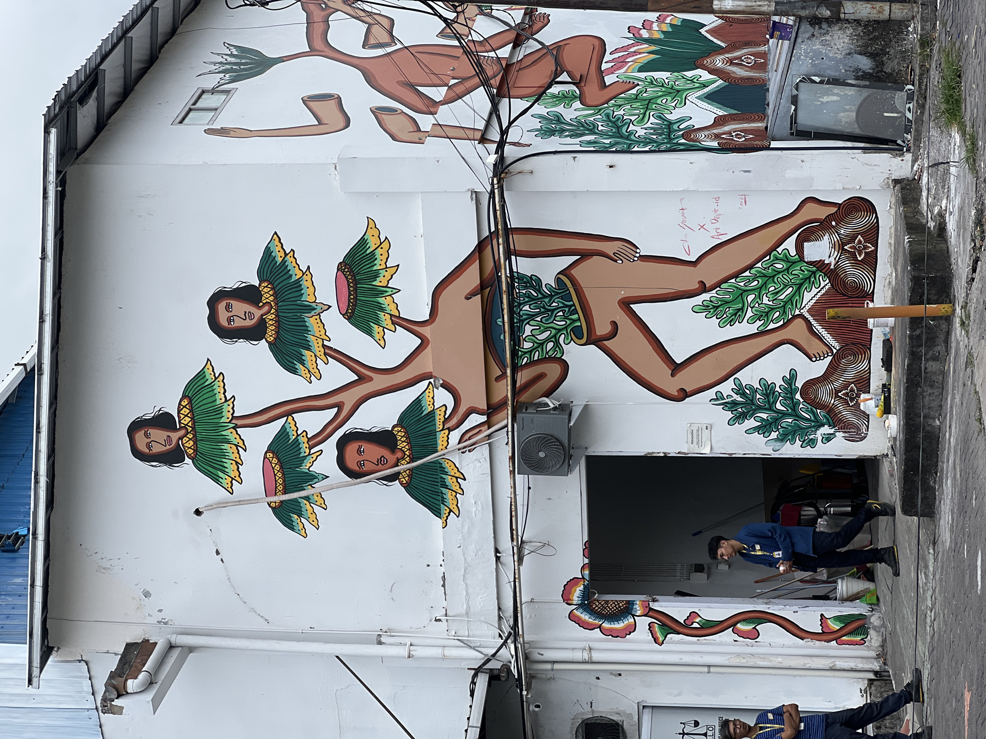 An eye-level, outdoor photograph captures the side of a building featuring a mural in Kuta, Indonesia. The mural is painted on a white, aged wall with visible cracks, depicting stylized human figures merged with floral or plant elements. One figure, located towards the top of the wall, is brown and appears to be in a prone position with dismembered limbs. Below this is a larger figure with three heads that are stylized as flowers with human faces at the center.
Two men are visible in the lower portion of the image. One is partially visible near the bottom, wearing a dark blue shirt, black pants, and glasses. The other man is in the middle of the image, walking through an open doorway. He is wearing a blue long-sleeved shirt and dark pants.
The building features an air conditioning unit mounted near the doorway. Wires run across the wall in front of the mural. The ground appears to be paved, and the overall environment suggests a bustling urban scene.