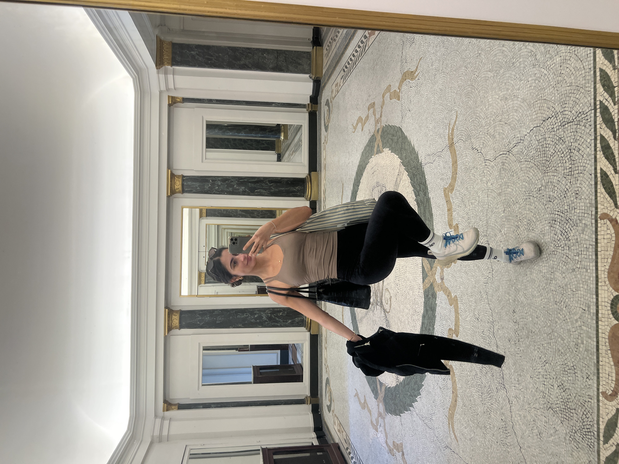 A young woman is captured in a selfie in an opulent indoor corridor, likely within a historic building in Paris, France. She is standing, looking towards the camera with a slight smile and making a peace sign with her left hand, which holds a smartphone. She wears a light brown tank top, black leggings, white socks with light blue accents, and white sneakers with light blue laces. Sunglasses are pushed up onto her head, and she carries a black purse and a striped tote bag over her shoulder. A black jacket is draped over her left arm.
The setting is distinguished by an intricate mosaic tile floor, featuring a large, ornate pattern in shades of white, grey, beige, brown, and green, centered around a circular design. The walls are adorned with large mirrors framed in white molding with gold accents. Between these mirrors are decorative elements resembling pilasters, featuring a dark green or black marble-like pattern with golden capitals and bases. These mirrored walls create an illusion of depth, reflecting the corridor and the woman multiple times. The ceiling is high, white, and features subtle molding, indicating a grand architectural style. The scene is well-lit, suggesting daytime. No discernible text is visible in the image.