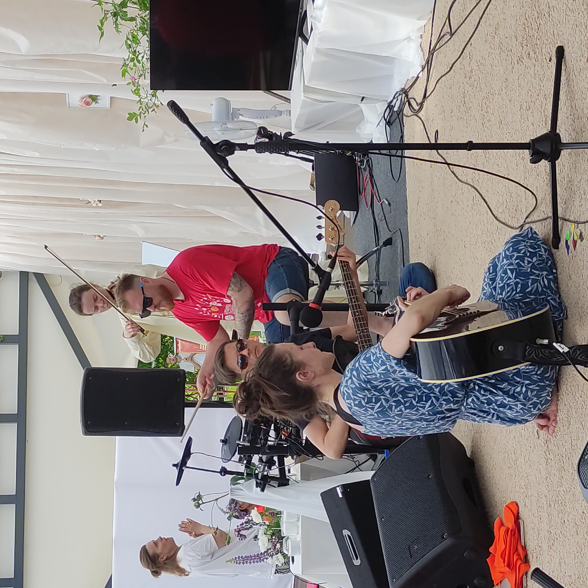 The image depicts an outdoor performance by a band in Zhirnosovo, Russia. In the foreground, a woman wearing a blue and white leaf-patterned dress is playing an acoustic guitar while seated on the ground. To her left is a black speaker. Behind her, a man wearing sunglasses and a red t-shirt is seated at a drum kit, holding drumsticks. To the left of the drummer, another man with sunglasses and a white t-shirt is visible. In the background, a woman in a white t-shirt is standing and gesturing with her hands, and another woman is playing the violin. A large white curtain is draped behind the performers. A black speaker is also visible to the far left of the image. The ground is covered with a light-colored carpet.