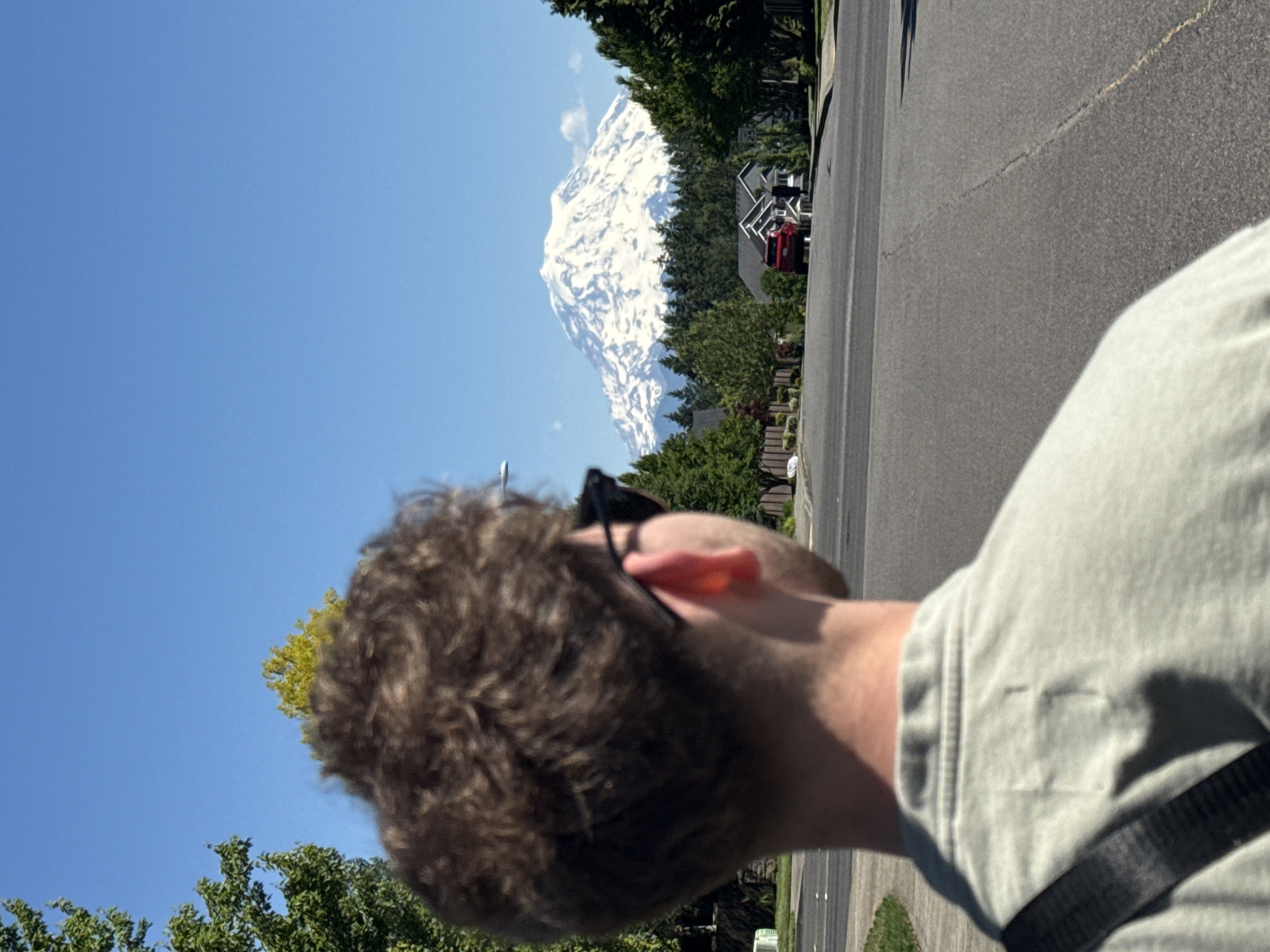 A medium shot captures the back of a young man wearing sunglasses and a light gray t-shirt, facing a snow-capped mountain range in South Hill, United States of America. The man's curly brown hair is visible, as is the strap of a dark bag draped over his shoulder. In the background, a paved road curves through a residential neighborhood lined with trees and houses. A red SUV is parked in front of one of the houses. The sky is clear and blue, and the sun is shining brightly, casting shadows on the road. The mountain range is majestic and imposing, dominating the horizon with its snow-covered peaks. The overall impression is one of a bright, sunny day in a peaceful suburban setting with a striking natural backdrop.