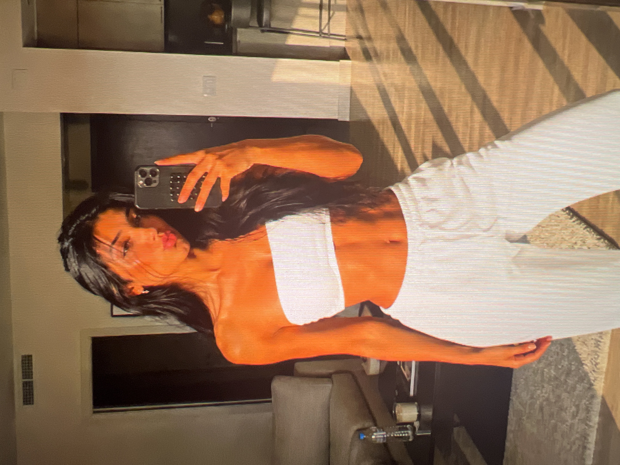 A young woman with dark hair and tanned skin is taking a mirror selfie in what appears to be an apartment. She is wearing a white bandeau top and white sweatpants. She is making a duck face at the camera and holding up her phone, which has a studded case. The background shows a living room area with a couch and a coffee table, and a kitchen area with a counter and stools. Sunlight is casting shadows across the wooden floor.