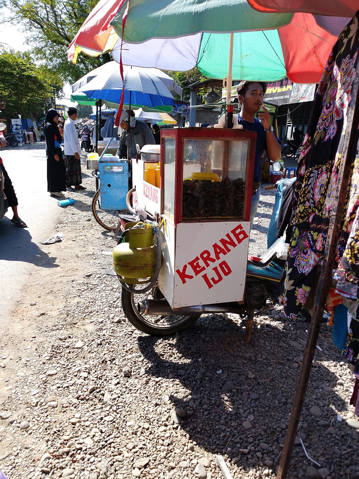 In Tegalwangi, Indonesia, a street vendor operates a mobile food cart under a canopy of umbrellas. The cart, which appears to be a modified motorbike, is filled with shellfish. A man, the vendor, is standing behind the cart, smoking. To the left of the cart, a woman in a black hijab and a man in a white shirt and sarong are standing, seemingly as customers. The ground is covered in gravel, and trees line the street, suggesting an outdoor, public setting. The scene is illuminated by sunlight, casting shadows on the ground.