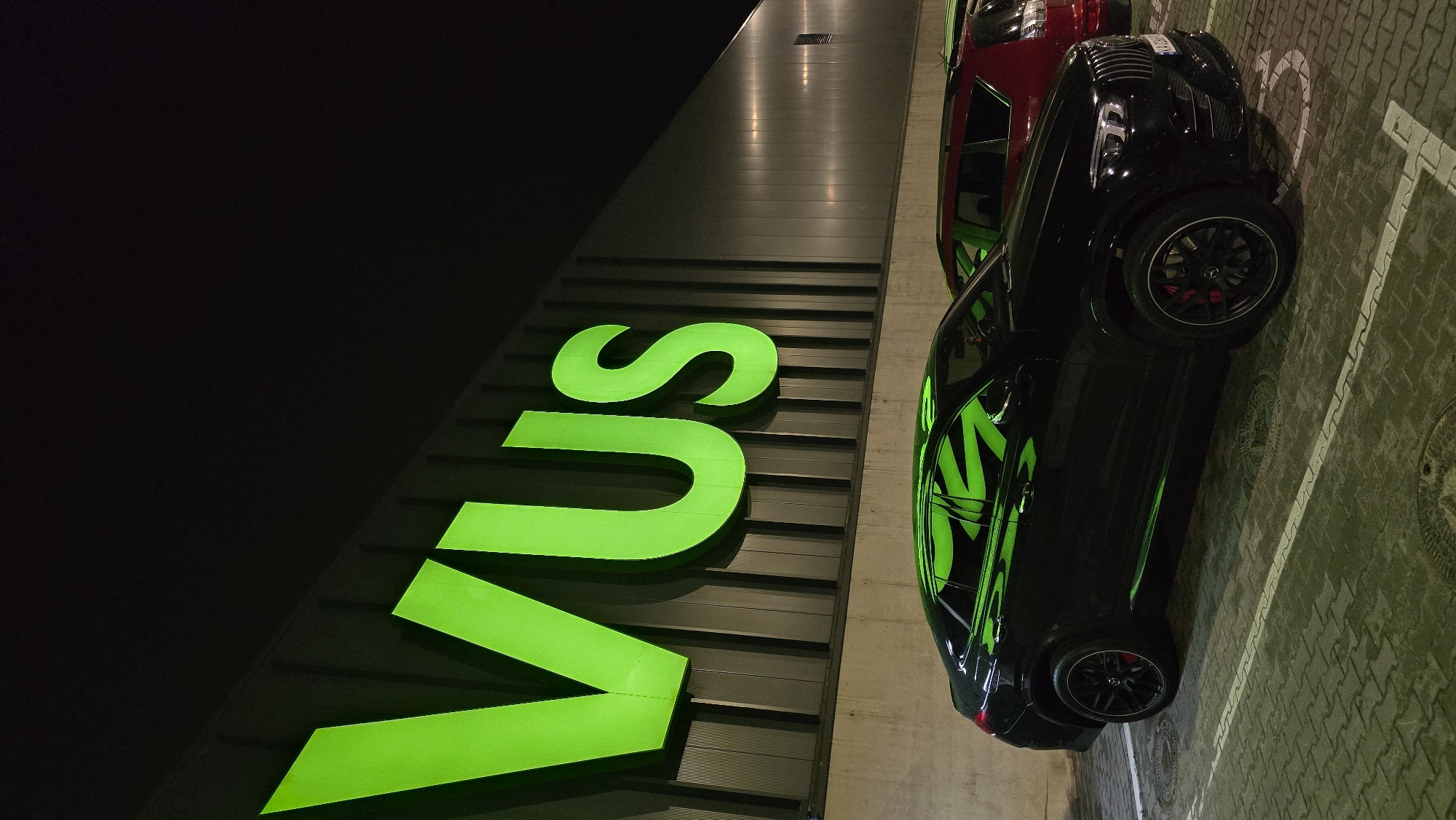 An overhead, tilted night shot captures a black high-performance SUV parked next to a building adorned with a large, illuminated green sign spelling "VUS". The dark setting, possibly a parking garage or outdoor lot at night, is characterized by the prominent green glow emanating from the sign, which strongly reflects off the glossy black paintwork of the SUV. The SUV features distinctive black multi-spoke wheels with bright red brake calipers, visible on the front left wheel. Behind the black SUV, a portion of a red vehicle, likely another SUV, is visible. The ground is paved with interlocking bricks, featuring faded white lines indicating parking spaces and several circular manhole covers. The top left corner of the image is completely dark, suggesting the vastness of the night sky or an unlit area of the structure. The overall mood is urban and modern, highlighted by the artificial lighting and sleek vehicles. No people are visible.