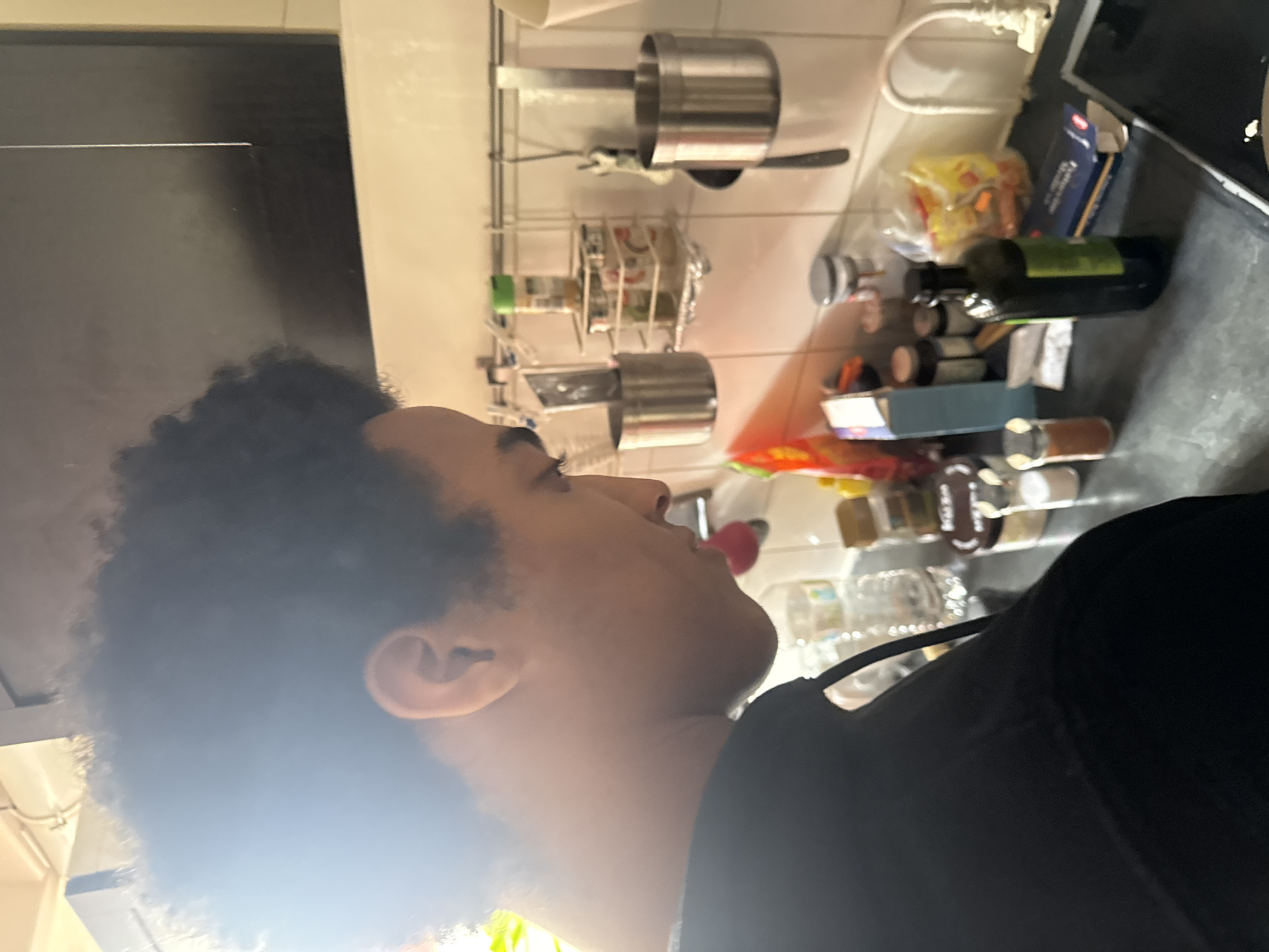 The image shows a person with dark skin and curly hair, wearing a black shirt, standing in a kitchen. The kitchen has white tiled walls and a counter with various objects, including spices, bottles, pasta, and a cookware rack mounted on the wall. The location of the image is not discernible as being in Paris, France.