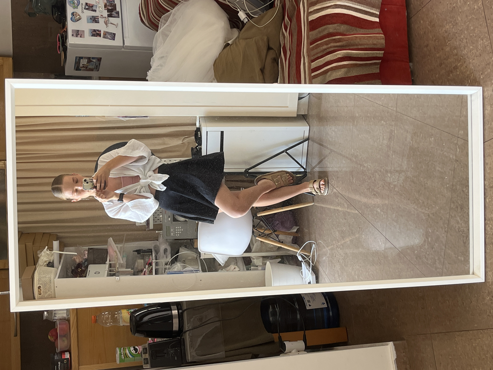 The image depicts a full-length mirror reflecting a young woman standing in what appears to be a cluttered residential room, potentially an apartment. The woman is wearing a white tied-front blouse over a pink camisole and a black skirt. She is holding a smartphone in her right hand and taking a selfie. She is wearing light brown sandals.
The background reflected in the mirror shows a chaotic environment with various objects. To the left, there are shelves filled with items including boxes, containers, and appliances like a black kettle and a toaster. A large blue water cooler is also visible. Further back, a white office chair is positioned in front of a tall white cabinet. To the right of the woman, a white refrigerator is covered with magnets and photos. A white tulle fabric, possibly part of a dress, is draped over a red and white striped couch.
The floor is tiled with large brown square tiles. The overall impression is one of a lived-in space with storage and household items present. The image does not contain any elements that would specifically indicate the city of Budva, Montenegro.