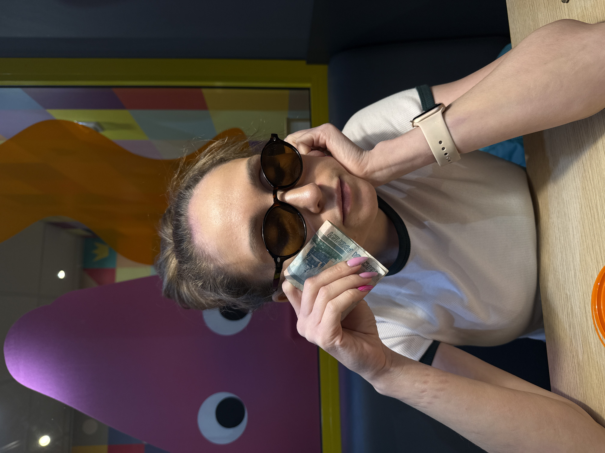 The image shows an adult female seated at a light wooden table inside a Dodo Pizza restaurant, likely located in Izhevsk, Russia. She has light skin, hair pulled back, and wears dark oval-shaped sunglasses. She is dressed in a white ribbed t-shirt with dark trim. A light beige smart watch is visible on her left wrist. Her right hand, adorned with pink manicured nails, some featuring a darker pink striped design, holds a folded Russian 1000 Ruble banknote near her face. Her left hand supports her head, resting on her chin, with her gaze directed downwards. She is seated on a dark blue upholstered booth. Behind her, a large window reveals a vibrant interior space. This background features a large pink cartoon blob-like character with two white, black-pupiled eyes, along with abstract orange shapes and a wall decorated with colorful geometric patterns in yellow, light blue, purple, and green. A yellow crown symbol is present within the patterned wall. Reflections of overhead lighting are visible in the window. The interior wall to the right is dark grey or navy blue.