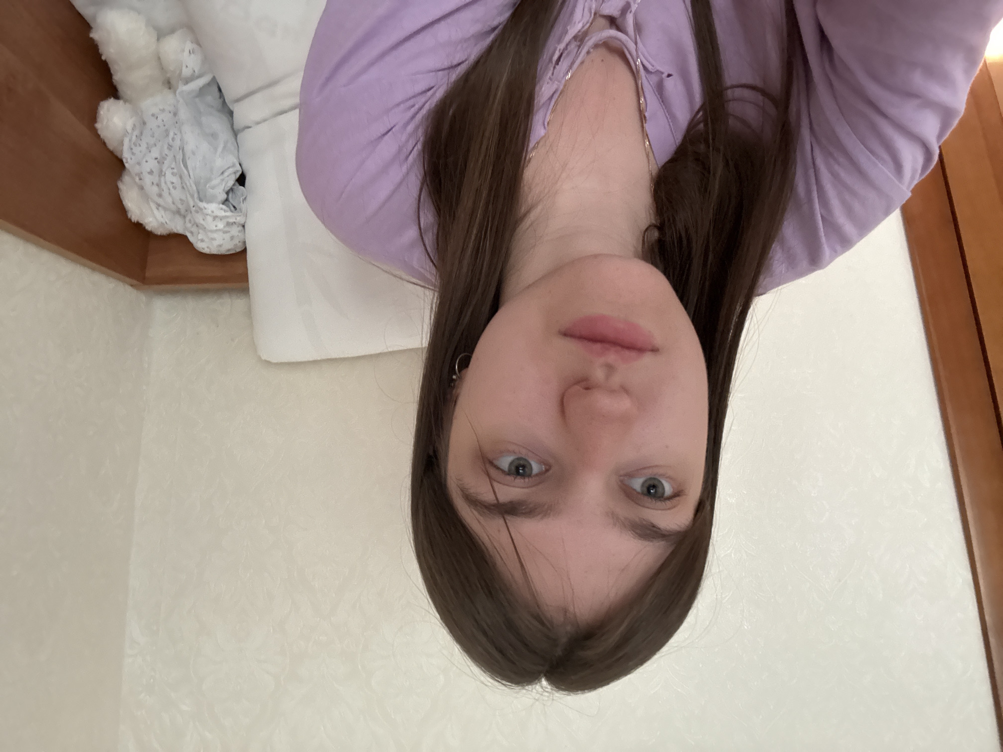 The image is an upside-down selfie of a fair-skinned young woman with long brown hair, wearing a light purple shirt. She has light blue eyes and visible earrings. The background includes a white pillow, a plush toy, a wooden headboard, and a patterned white wall. A wooden doorframe is also visible on the right side of the image. The location is unidentified.