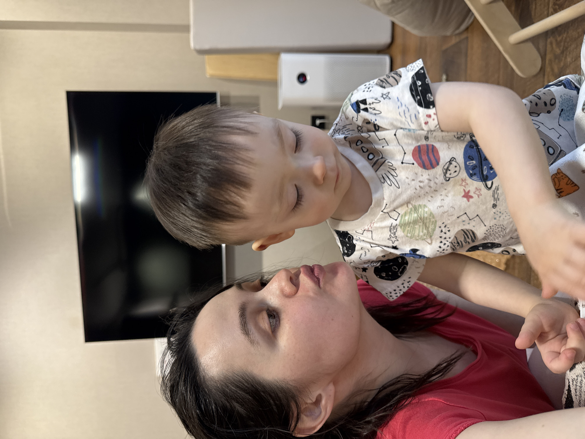 This image captures a tender, intimate moment indoors between an adult woman and a young child. The perspective is a Dutch angle, with the subjects oriented diagonally across the frame.
The woman, with dark hair, is lying down, wearing a red top. Her head is slightly raised as she looks up, her gaze fixed on the child above her, conveying a sense of tenderness or deep focus. The child, a toddler or preschooler with short dark hair, is leaning over the woman, looking down at her with eyes nearly closed, suggesting a moment of quiet contemplation, affection, or sleepiness. The child wears a white t-shirt patterned with colorful space-themed illustrations, including planets and rockets.
In the background, a large, dark television screen is visible on a light-colored wall, indicating it is turned off. To the right of the television, on what appears to be a light wooden surface, rests a white rectangular air purifier or humidifier, displaying a small red indicator light. Partially visible on the far right is a piece of light-colored wooden furniture, possibly part of a child's high chair or play equipment. The flooring appears to be wooden. The setting is a cozy indoor environment, likely a home in Izhevsk, Russia. There is no discernible text visible in the image.