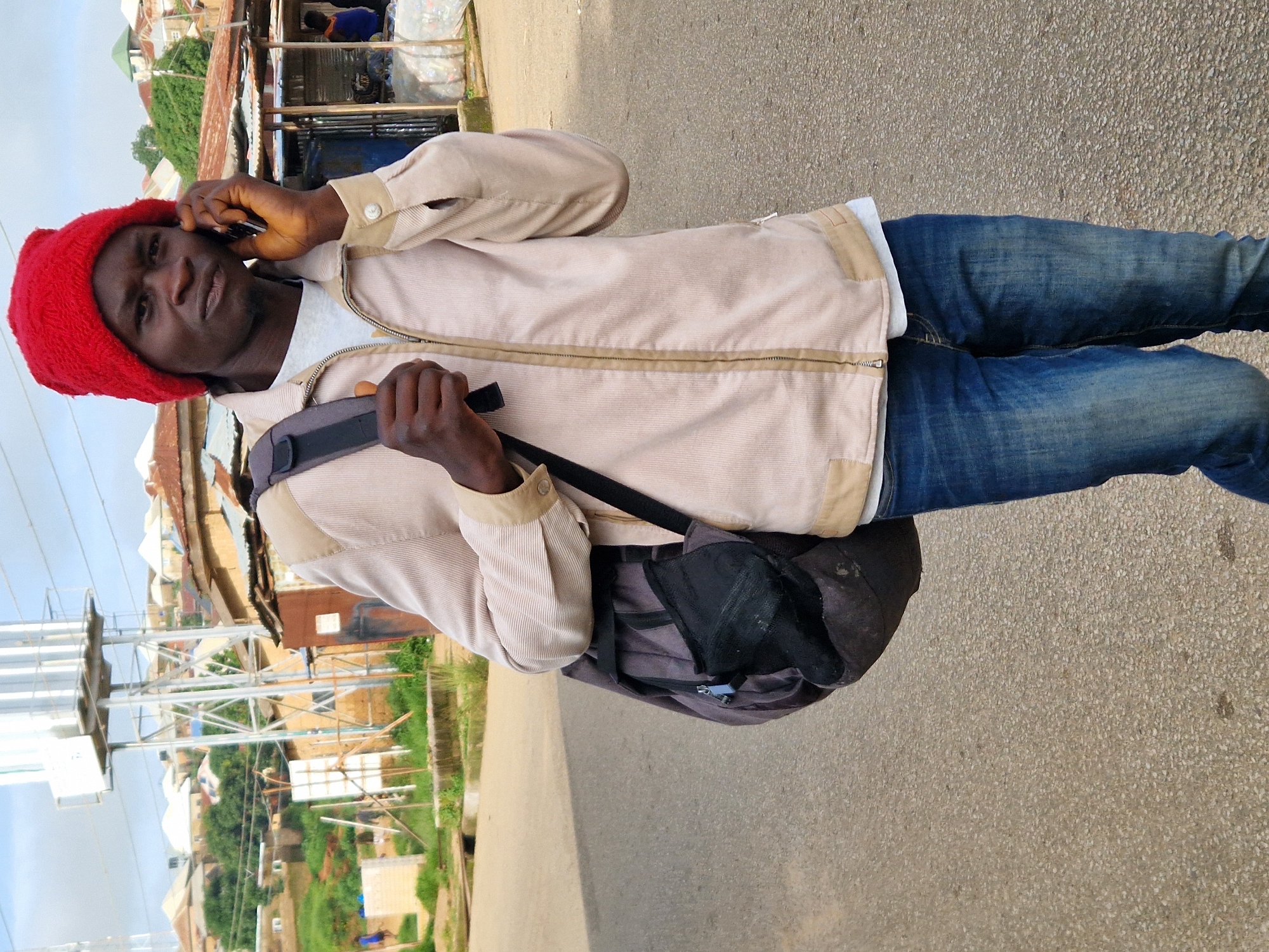 A man wearing a red beanie and a tan jacket is standing on a paved road in Kabong, Nigeria, and talking on a mobile phone. He has a dark backpack slung over his shoulder. In the background, there are buildings with corrugated metal roofs, a tall water tower, and some trees. The sky is overcast.