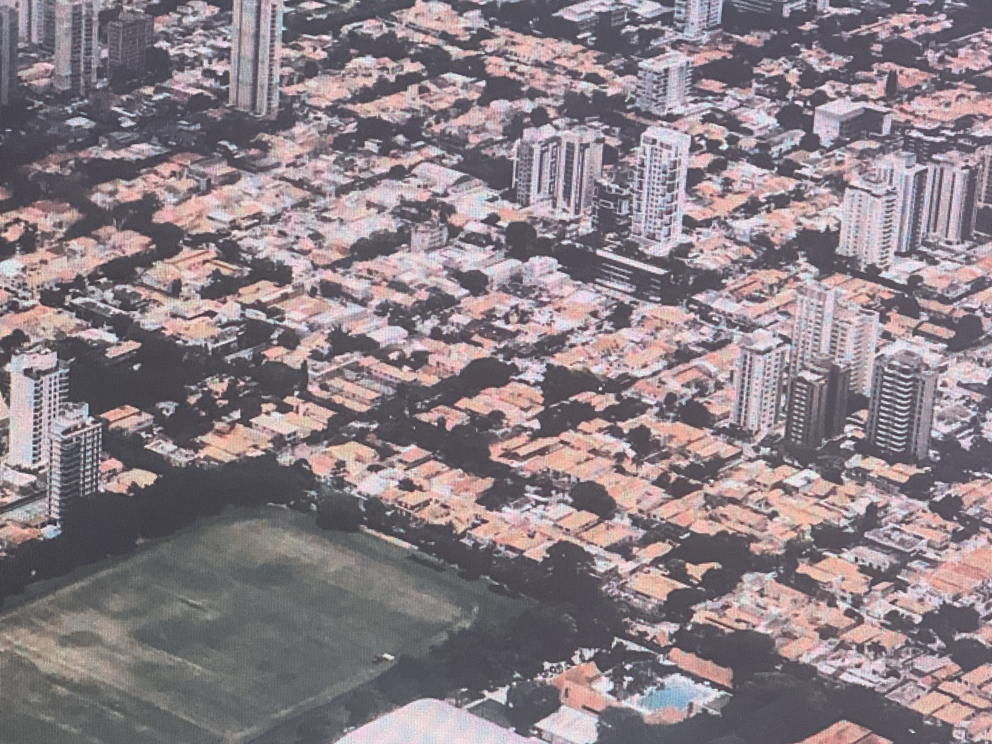 An aerial view of a densely populated urban area showcases a mix of high-rise buildings and lower-rise residential structures with reddish-orange tiled roofs. The buildings are tightly packed, interspersed with dark green foliage, suggesting the presence of trees and landscaping. A large, open green space, possibly a park or sports field, occupies the lower left portion of the image, with some visible markings or pathways. The overall scene depicts a developed urban environment with varying architectural styles and land use.