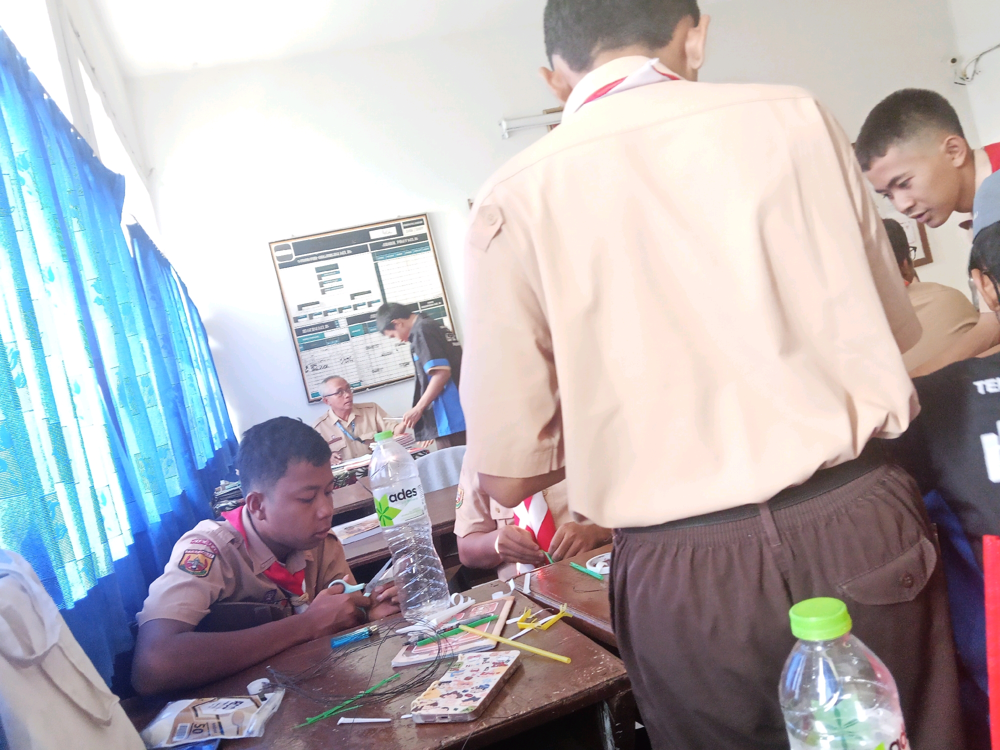 The image depicts a classroom scene in Mangunjaya, Indonesia, likely a school due to the presence of students in uniforms and educational materials. In the foreground, a young male student in a tan uniform with a red scarf is seated at a wooden desk, using scissors to cut green string. His focus is on the task at hand. Beside him on the desk, a clear plastic bottle of "Ades" brand water stands upright, next to various crafting materials including colored straws, wires, and what appears to be a decorated phone case.
In the background, several other individuals are visible. To the left, a whiteboard displays charts and text, suggesting an academic lesson is taking place or has recently concluded. Another student in a tan uniform is partially visible in the center background. To the right, a male student with a shorter haircut is leaning forward, observing something on a desk. Further back, an adult male in a tan uniform with glasses is seated, also appearing to be an instructor or supervisor. The room is lit by natural light, possibly from a window covered by blue curtains on the left, and artificial lighting from above. The overall atmosphere appears to be one of focused activity and learning.