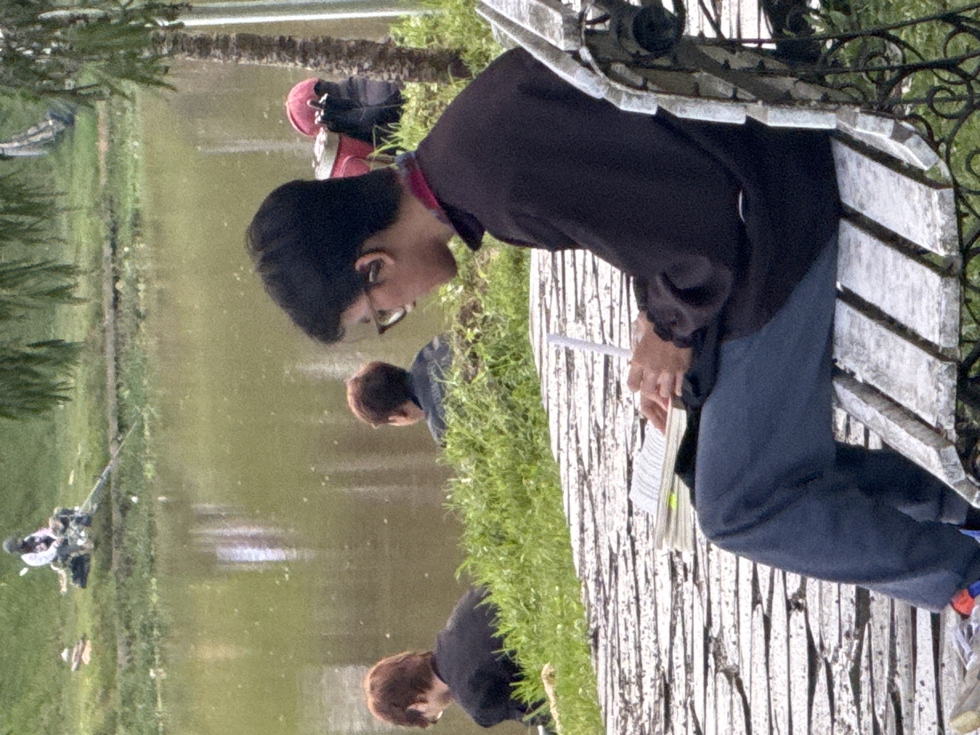 The image depicts a man sitting on a white, ornate park bench in Da Lat, Vietnam. The man is wearing glasses, a black beanie, a dark brown long-sleeved shirt, and blue pants. He is reading a document or book. Behind him, there is a lake with fishermen along the shoreline. There are two other individuals partially visible standing on the grassy bank near the bench. The scenery includes trees and vegetation typical of the Da Lat region.