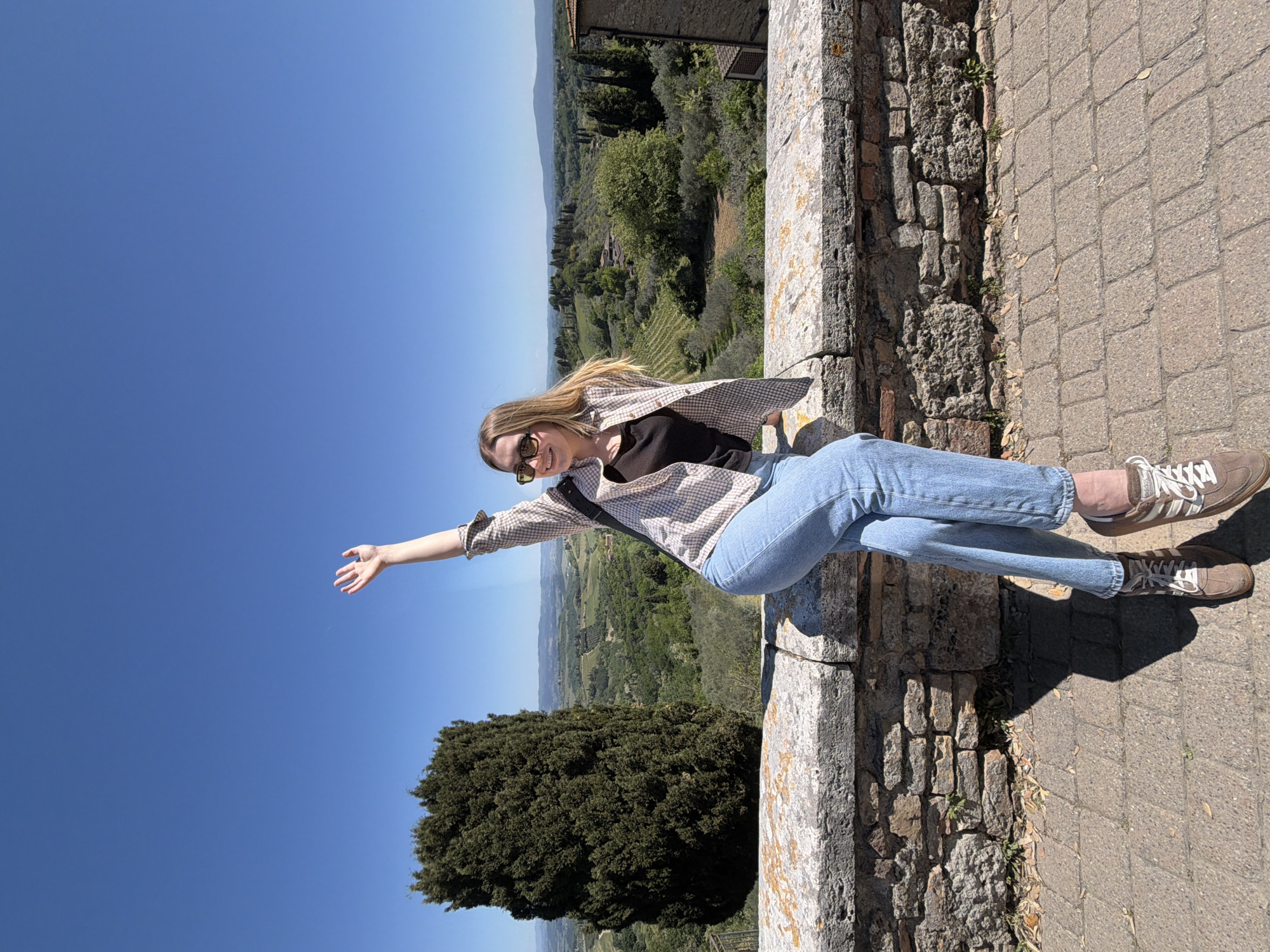 A full-length, rotated photograph captures a young woman smiling and posing on a stone wall, overlooking the scenic Tuscan countryside of San Gimignano, Italy. The woman has long blonde hair, wears sunglasses, a light-colored plaid shirt over a dark top, blue jeans, and brown sneakers with white laces. She is positioned horizontally in the frame, with her left arm outstretched as if reaching towards the distant landscape, and her body leaning back against the stone wall. Her bright, open smile conveys a sense of joy and relaxation, suggesting she is enjoying the moment and the view.
Behind her, the stunning backdrop features rolling green hills, lush vineyards, and numerous trees, including a prominent dark green conifer in the mid-ground. Part of a historic stone building or structure is visible in the upper background, characteristic of the region. The sky is a vast, clear blue, indicating bright, sunny weather. Strong shadows on the paved path and wall suggest the image was taken during midday or early afternoon. The immediate foreground includes the textured, ancient-looking stone wall she is resting on and a section of paved path. The unique rotation of the image gives a playful, almost gravity-defying feel to her pose, capturing a memorable moment amidst the beautiful Italian landscape. There is no visible text in the image.